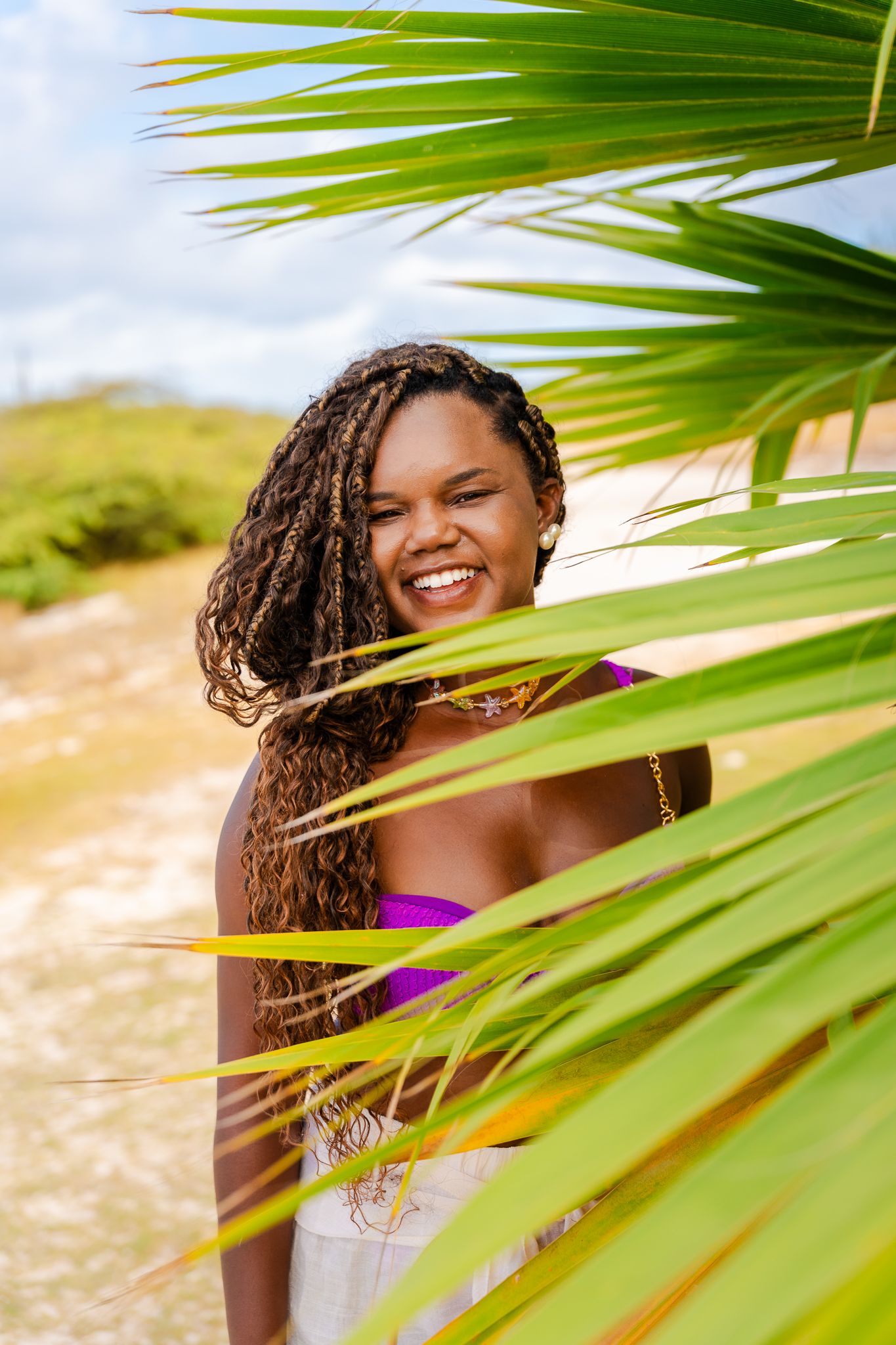 Smiling woman with braids in purple bikini top posing behind green palm fronds on a sunny beach.