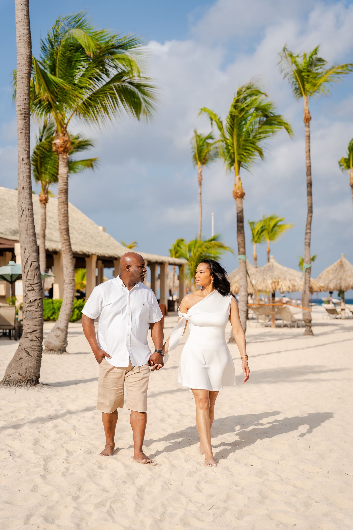 A couple walks hand-in-hand on a white sandy beach with palm trees, the woman in a white dress and man in casual attire.