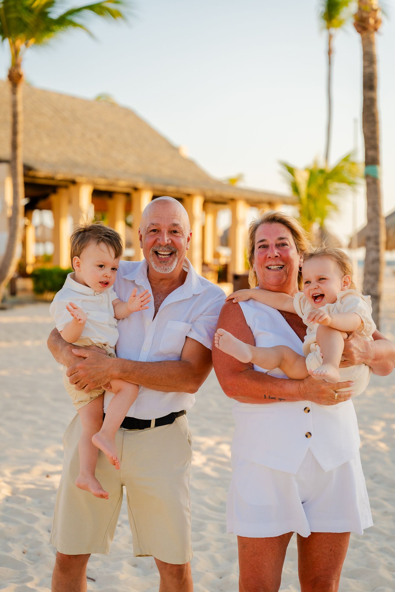 Grandparents laughing on a tropical beach holding two smiling babies, with palm trees and a thatched hut in the background.