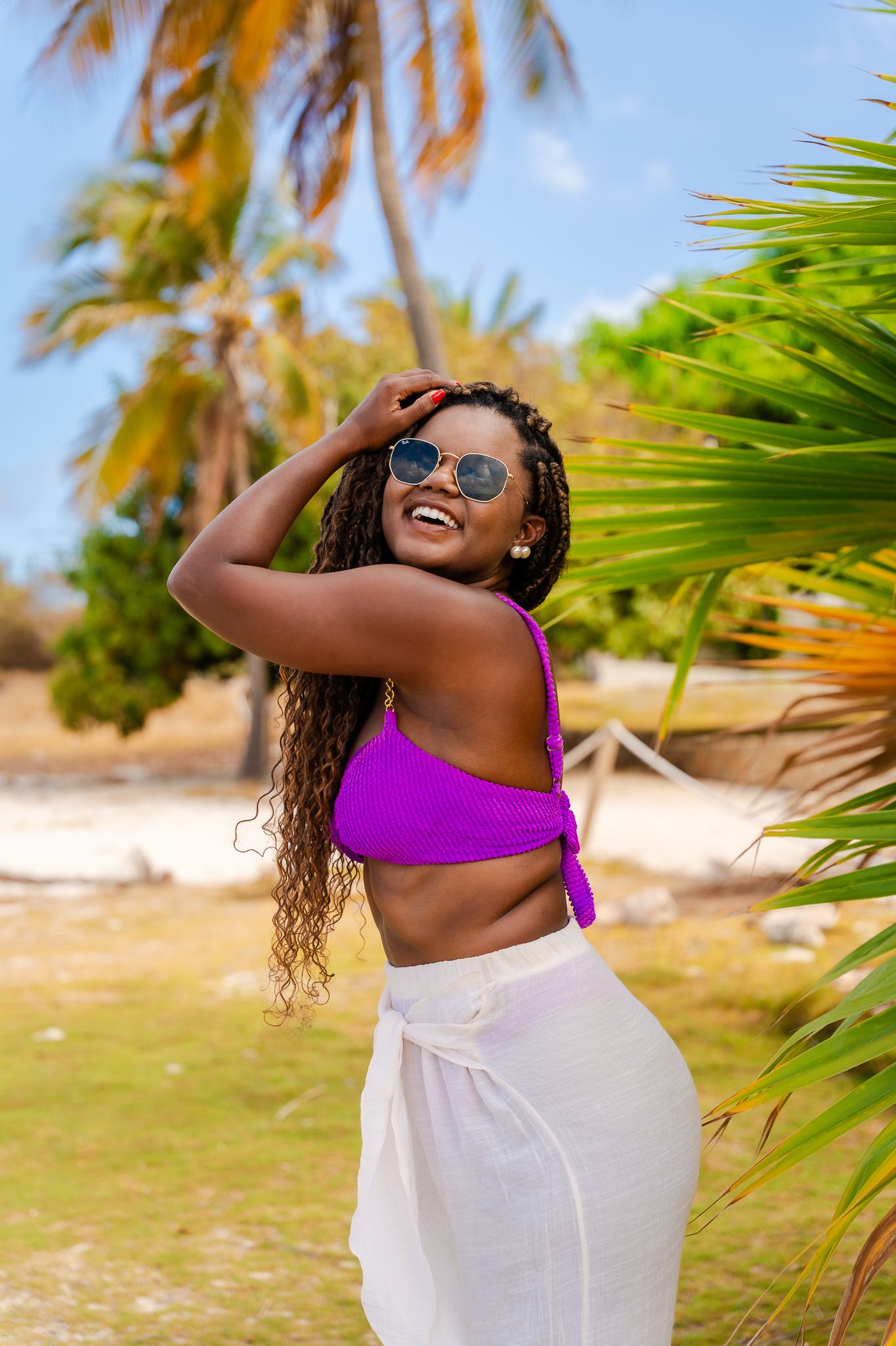 Smiling woman in purple crop top and white pants posing near palm trees on a sunny tropical beach.