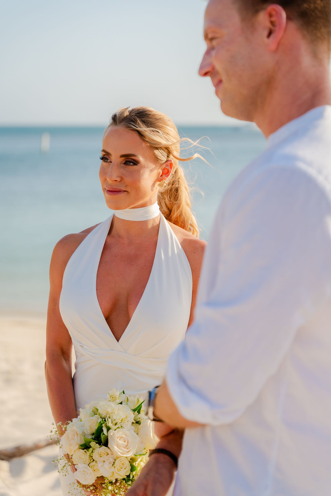 Bride in white halter dress holding bouquet faces groom on sandy beach with turquoise ocean backdrop.