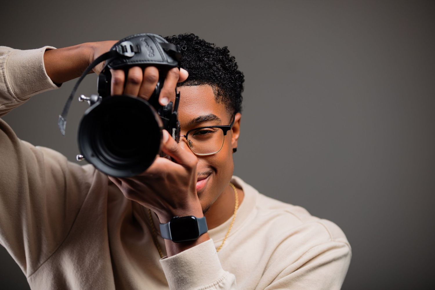 Young Black photographer in glasses and cream sweater taking a photo with a DSLR camera against gray background.