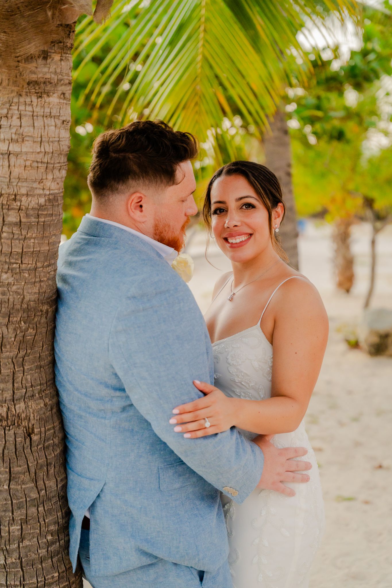 A smiling bride in a white gown embraces a groom in a light blue suit near a palm tree on a tropical beach.