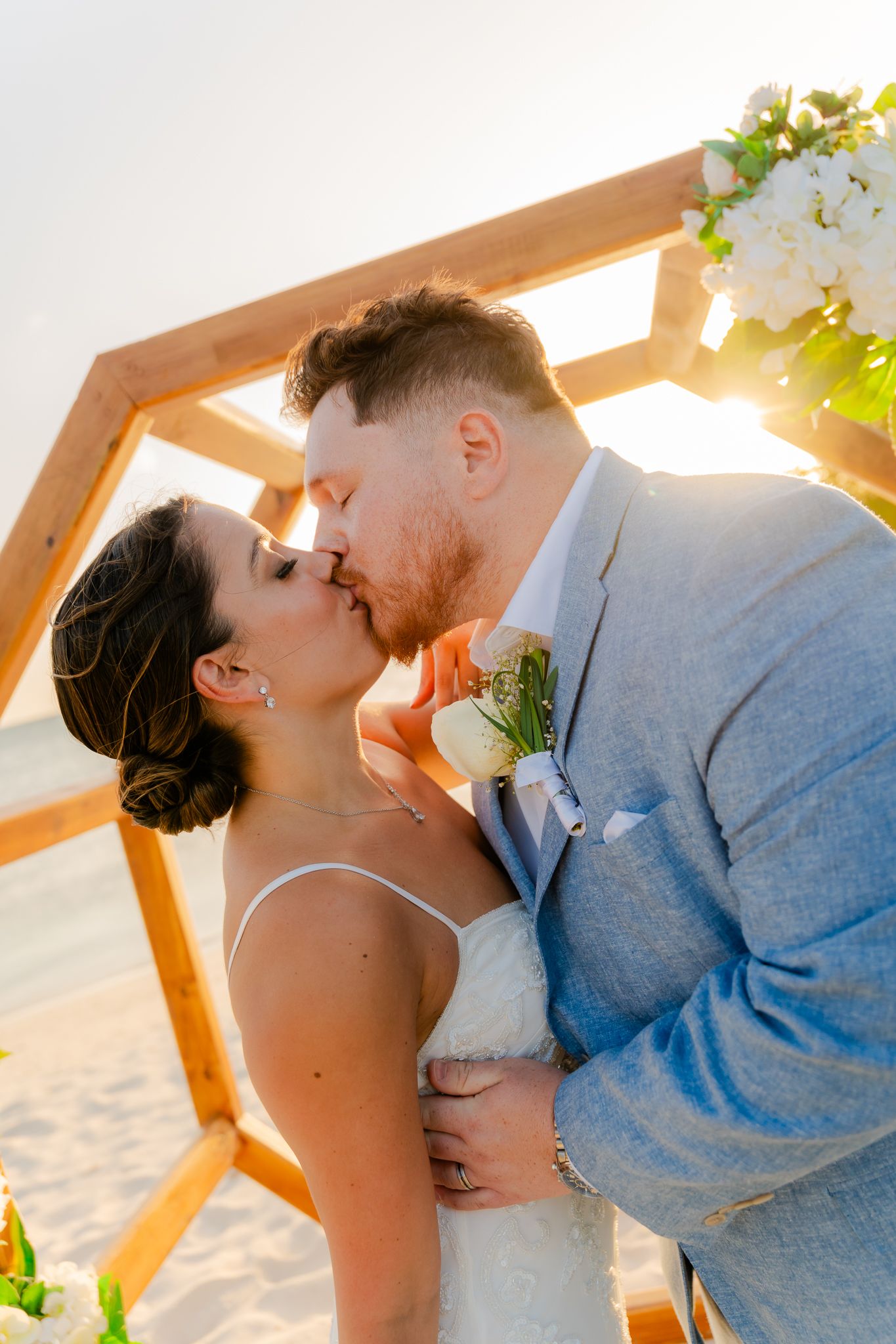 Bride and groom share first kiss under wooden arch on sunny beach wedding ceremony.