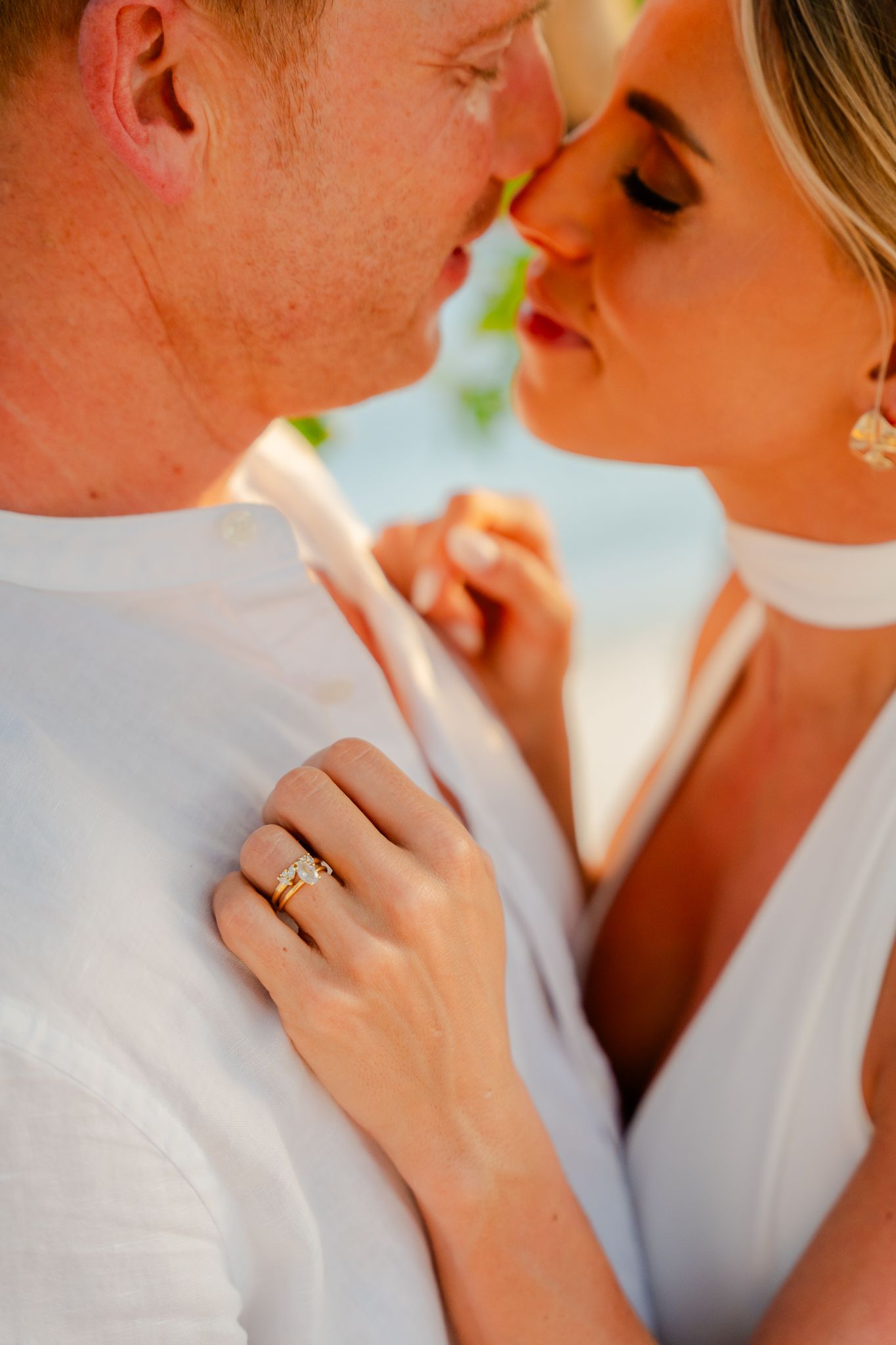 Couple in white outfits sharing a romantic moment, woman wearing gold rings, soft warm sunset light.