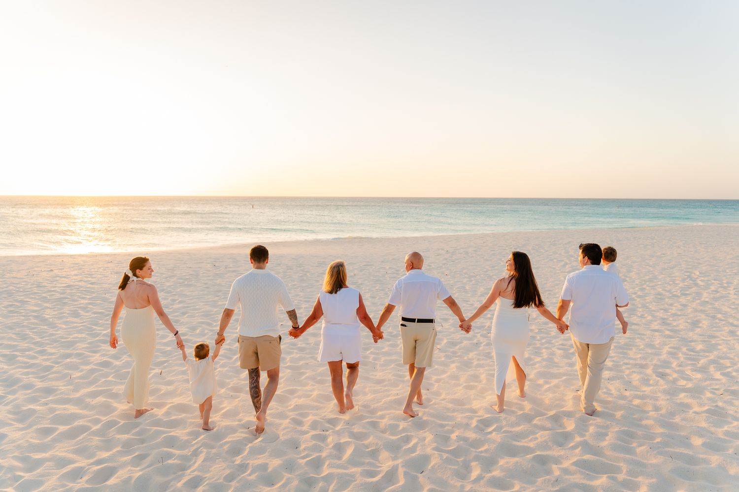 Family holding hands walking on a white sand beach at sunset wearing coordinated white outfits.
