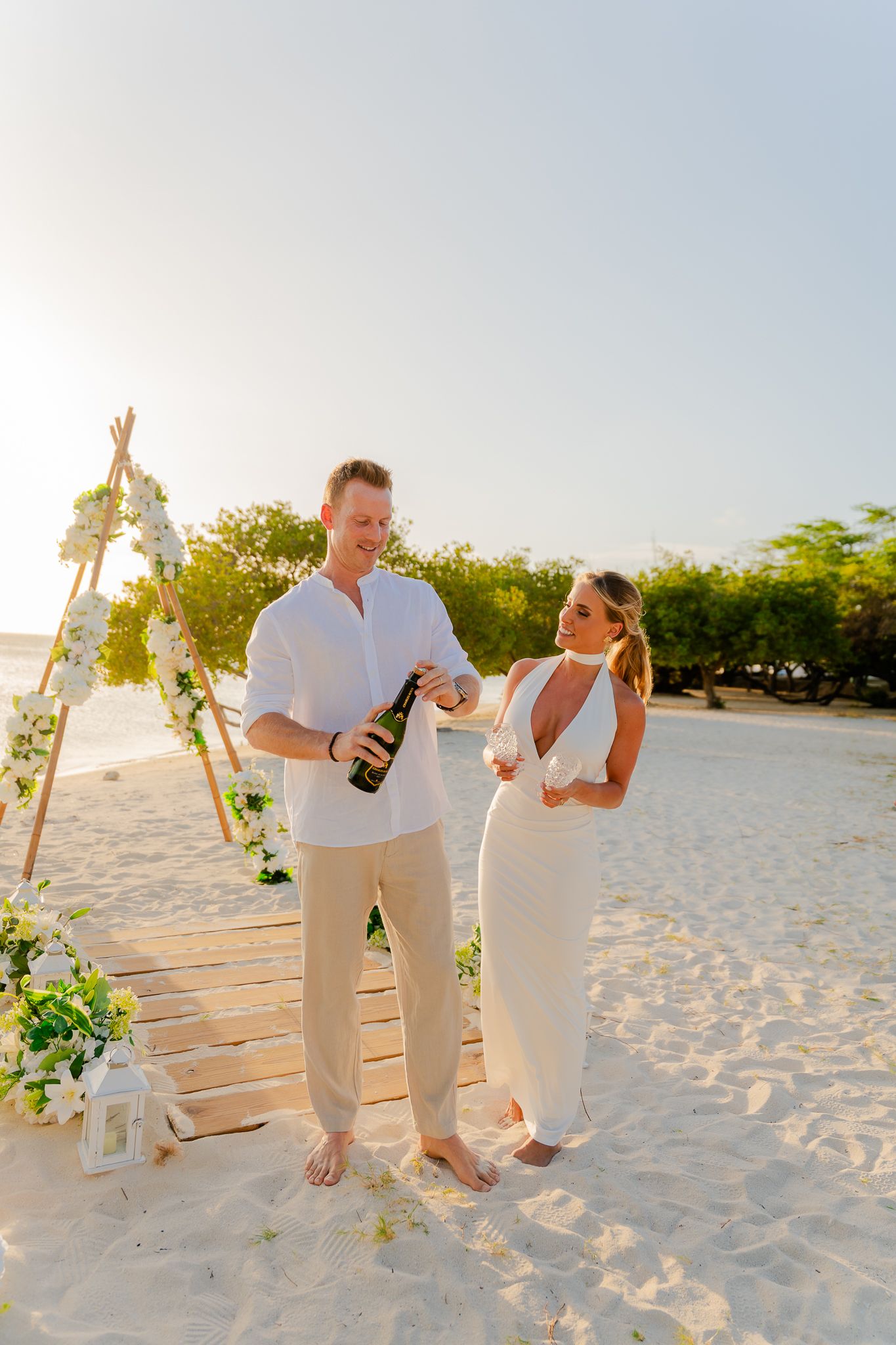 Couple in white outfits celebrating on a sandy beach with a floral wooden arch at sunset, opening champagne.