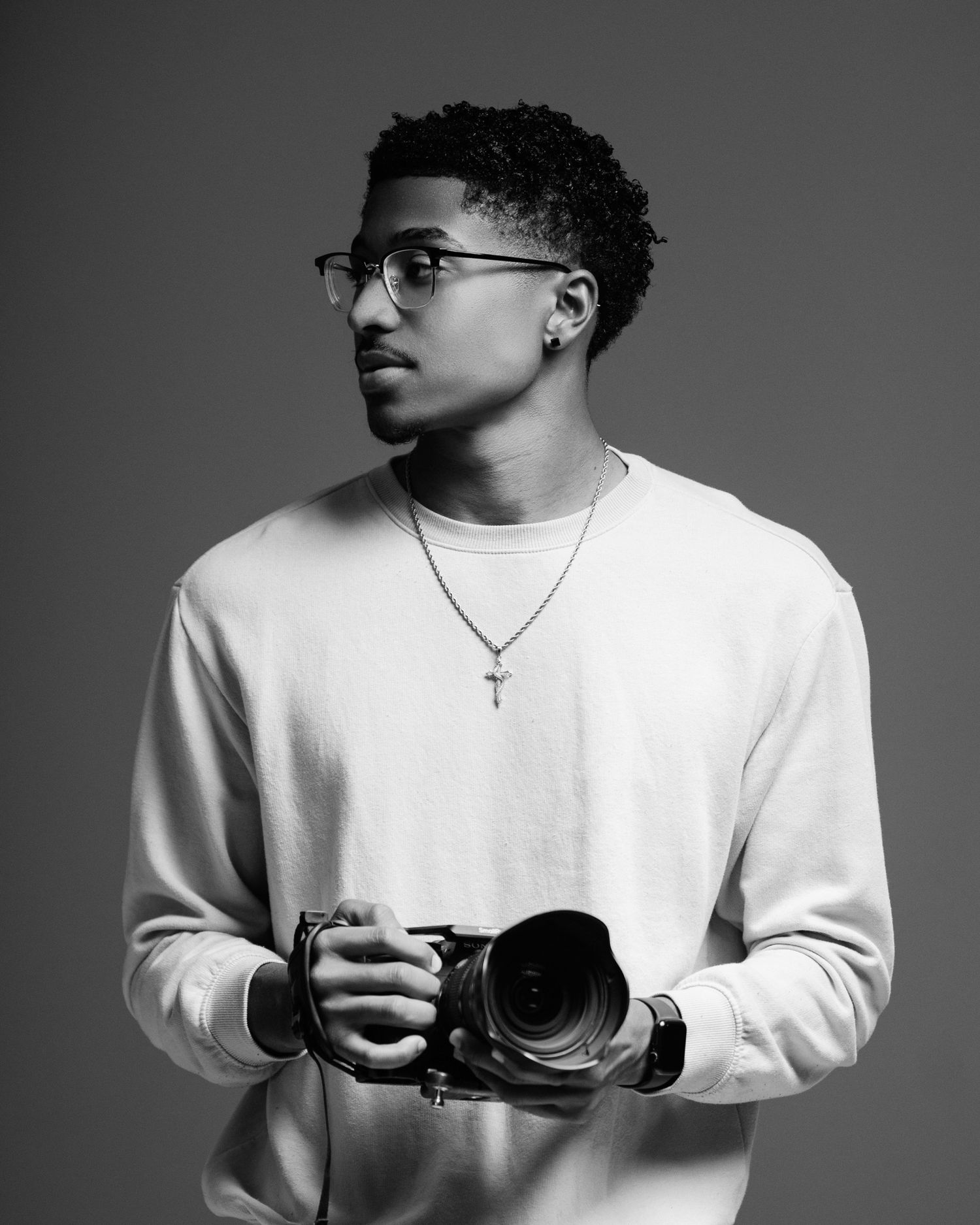 Young man with glasses holding a camera, wearing a white sweatshirt and cross necklace, black and white portrait.