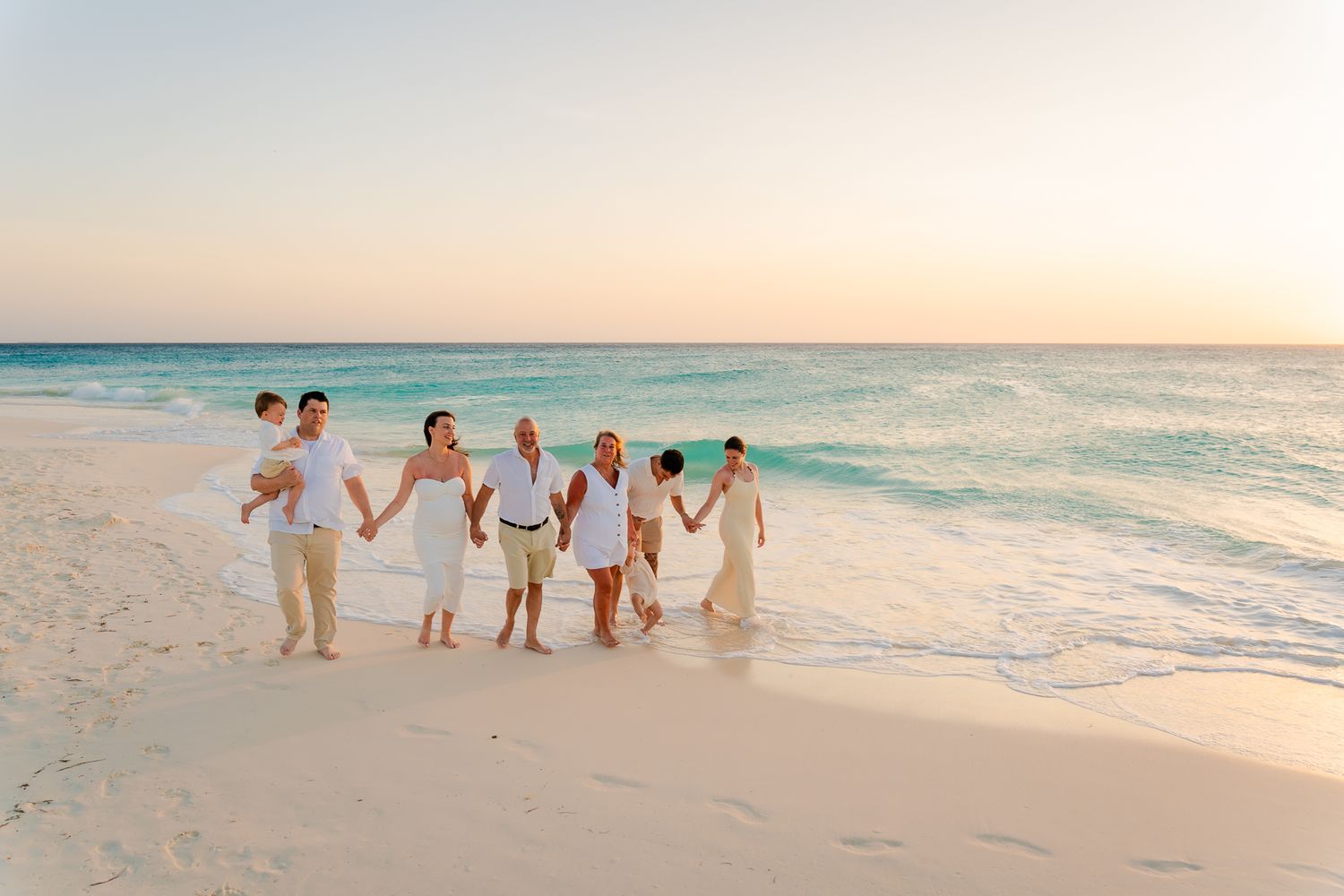 Family group in white outfits walking hand-in-hand on a tropical beach at sunset with turquoise waters.