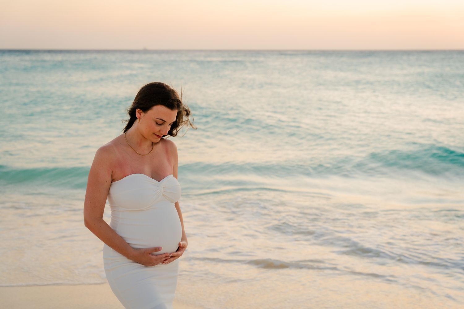 Pregnant woman in white dress cradling her baby bump on a serene beach at sunset.