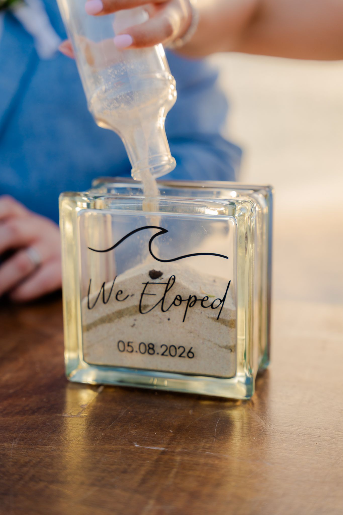 Sand being poured into a glass block engraved with We Eloped and date 05.08.2026 at a beach elopement ceremony.