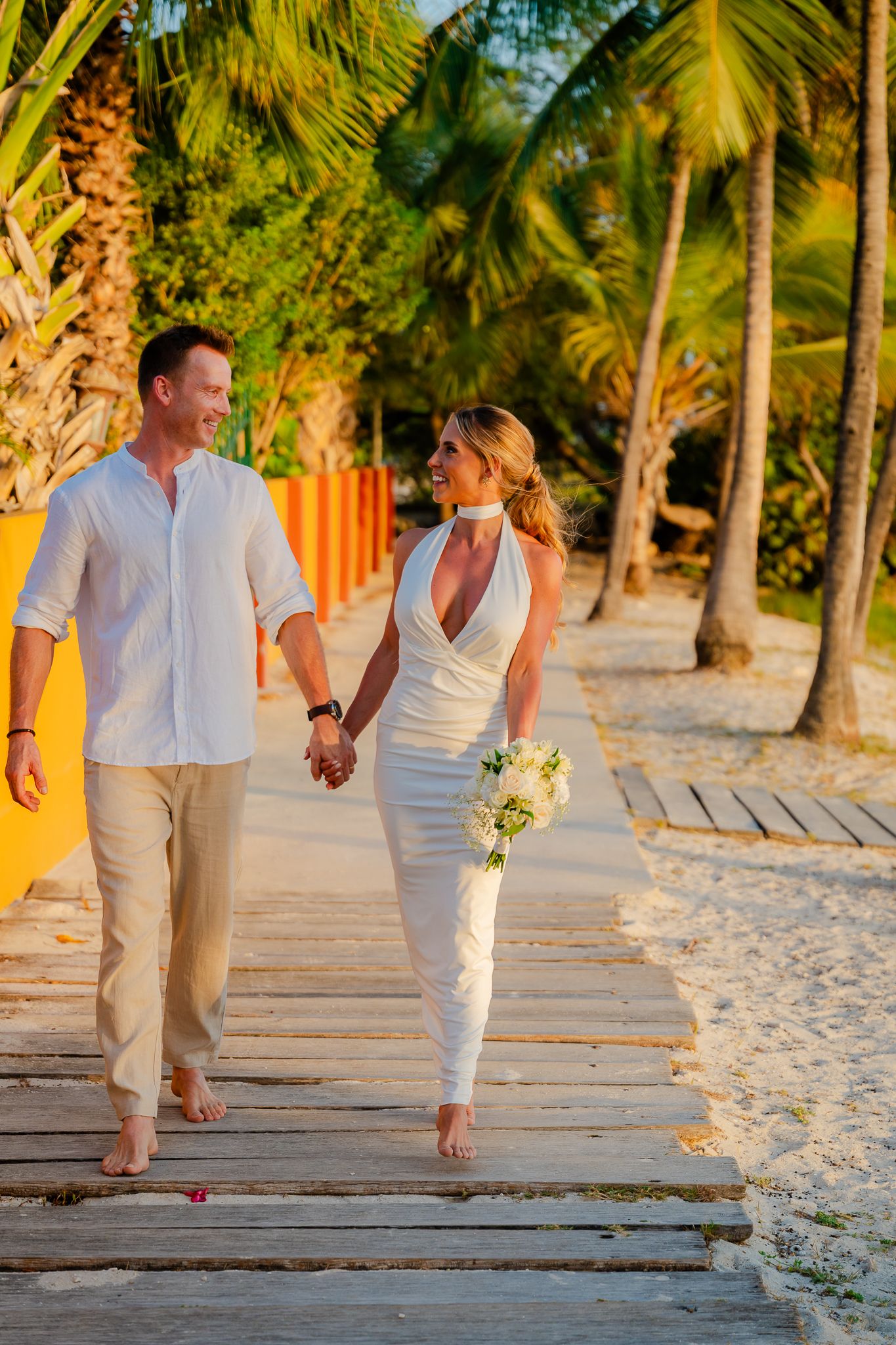 Couple in white wedding attire holding hands walking on a wooden boardwalk surrounded by palm trees on a tropical beach.