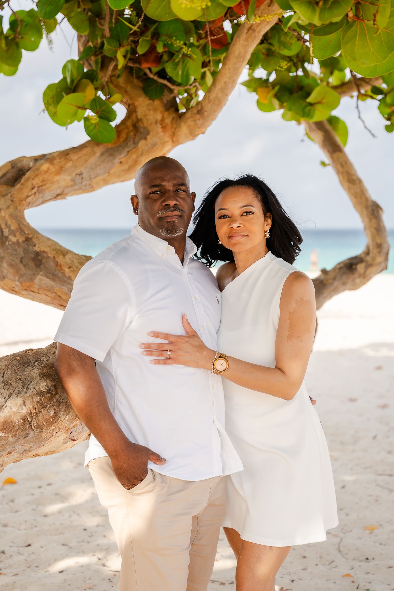 A couple in white outfits poses under a tropical tree on a sandy beach with turquoise water in the background.
