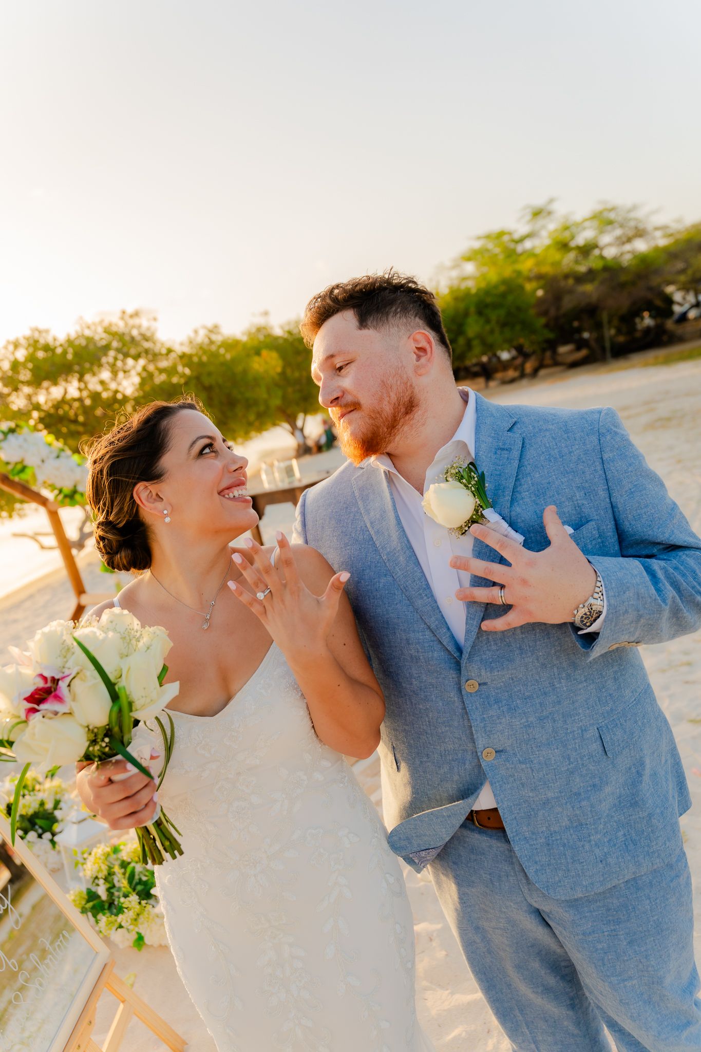 Bride and groom laughing together on beach, she shows ring in white dress, he wears light blue suit with boutonniere.