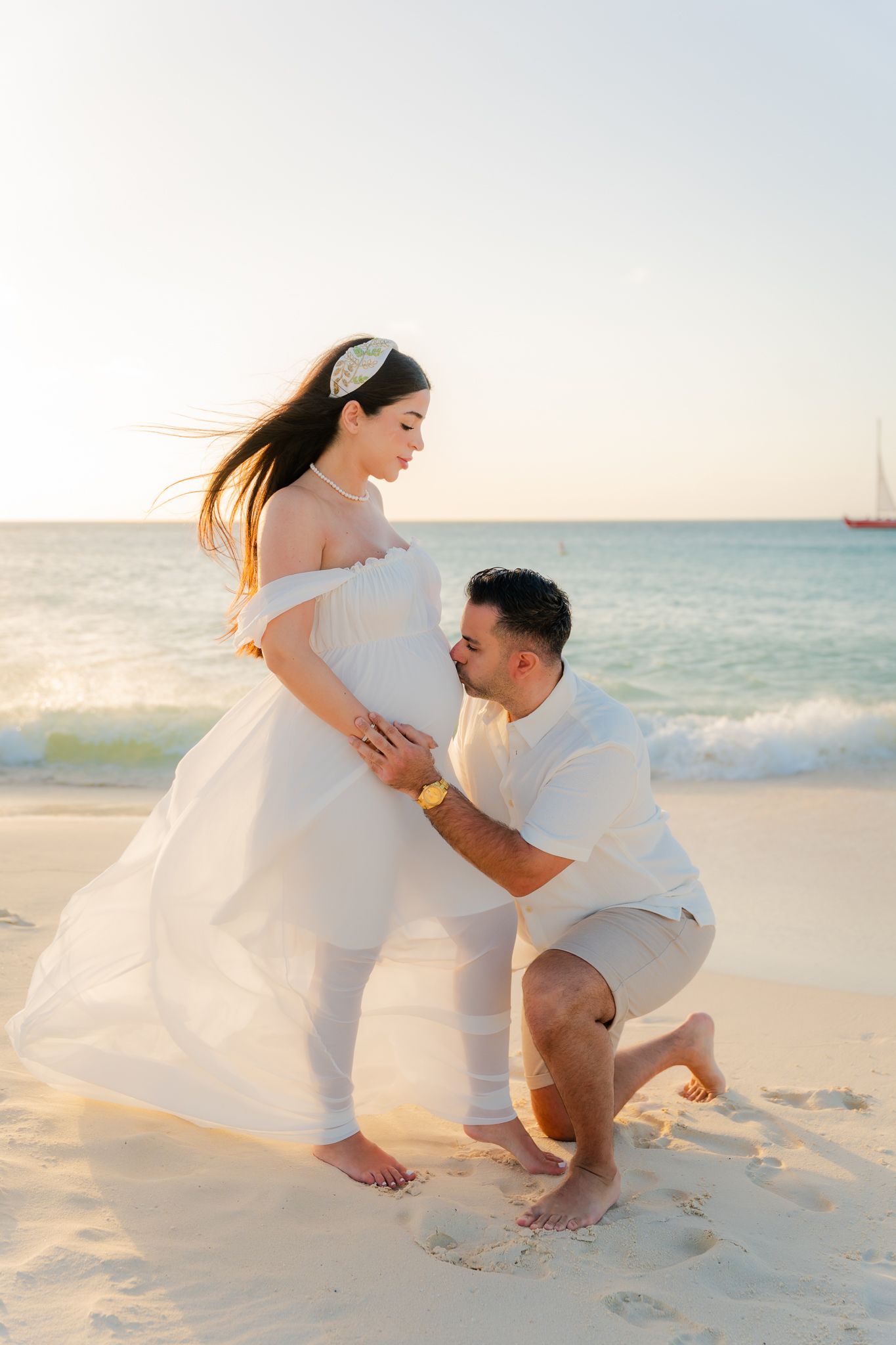 A man kneels on a sandy beach holding a pregnant woman in a white flowing dress during a golden sunset maternity photoshoot.