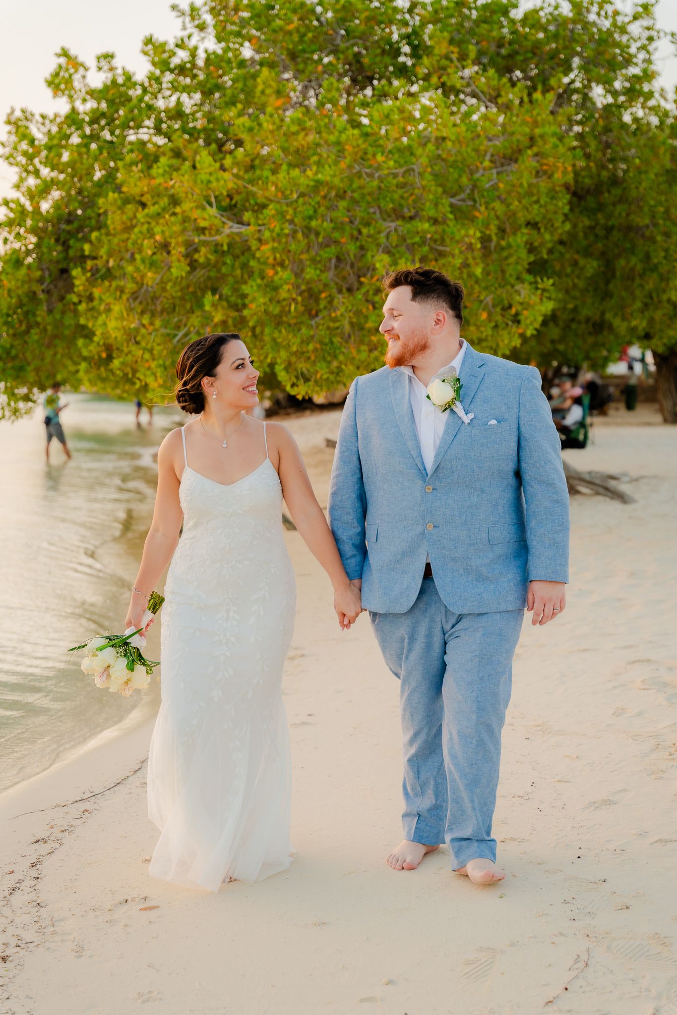 Happy newlyweds walk hand in hand barefoot on tropical beach at golden hour sunset.