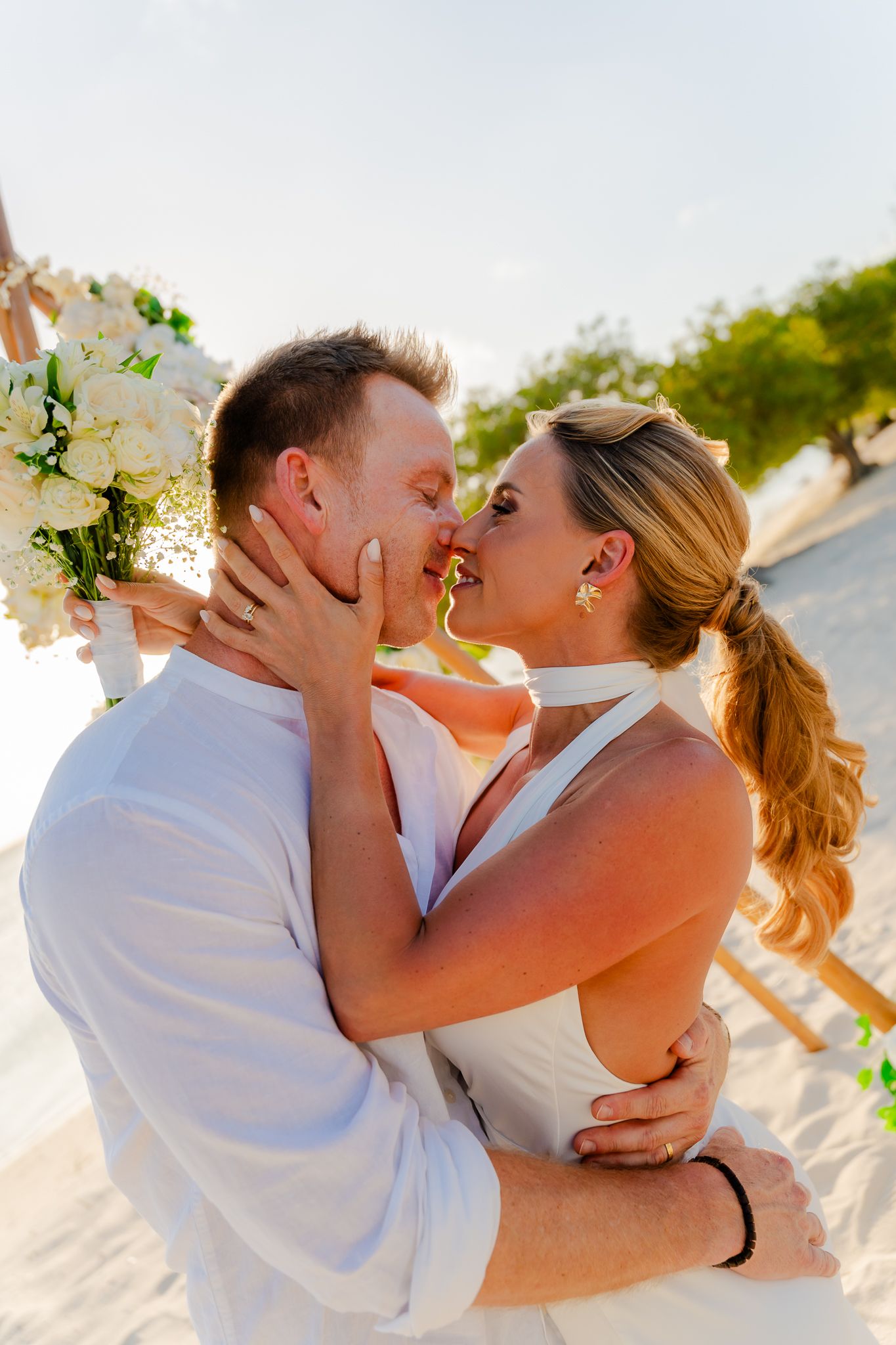 Newlywed couple sharing a romantic kiss on a beach wedding, bride holding white bouquet in a white dress.