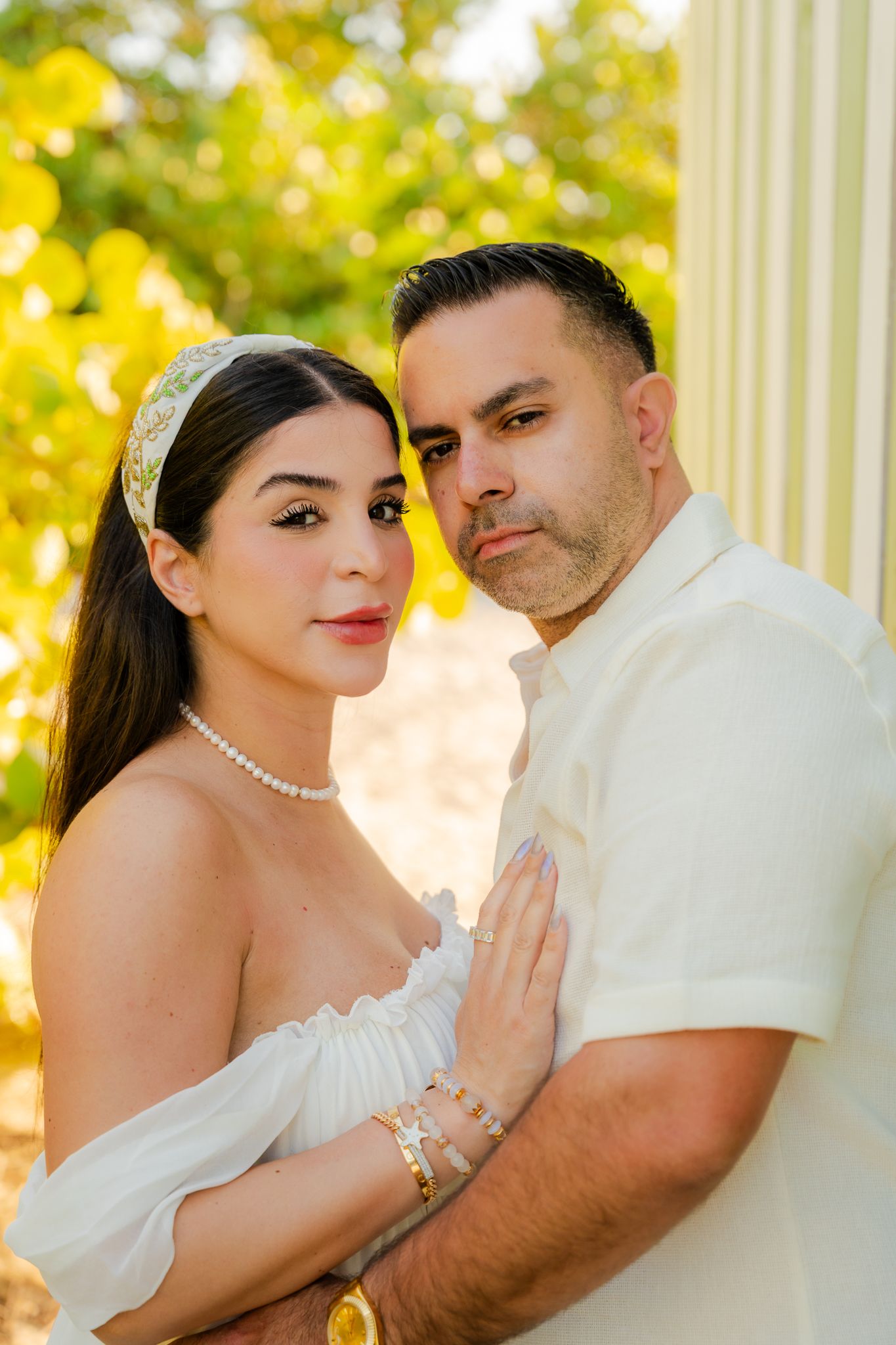 A couple in white attire poses elegantly outdoors, the woman wearing a pearl necklace and floral headband.