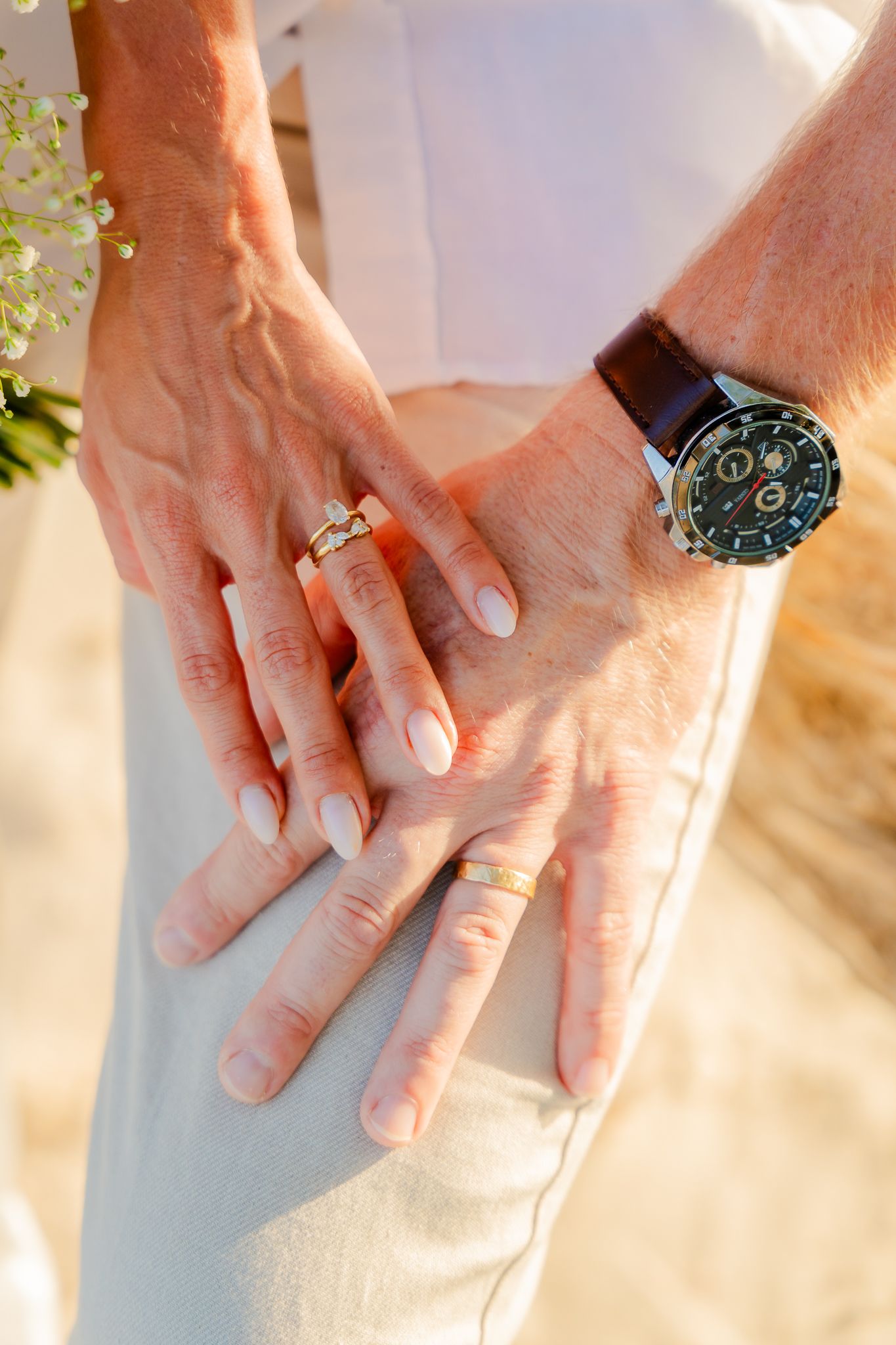 Close-up of couple's hands showing wedding rings and a brown leather watch during a beach wedding ceremony.