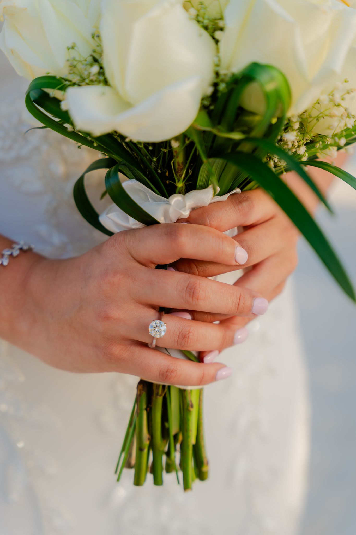 Bride holding a white tulip bouquet with a round diamond engagement ring and delicate bracelet visible.