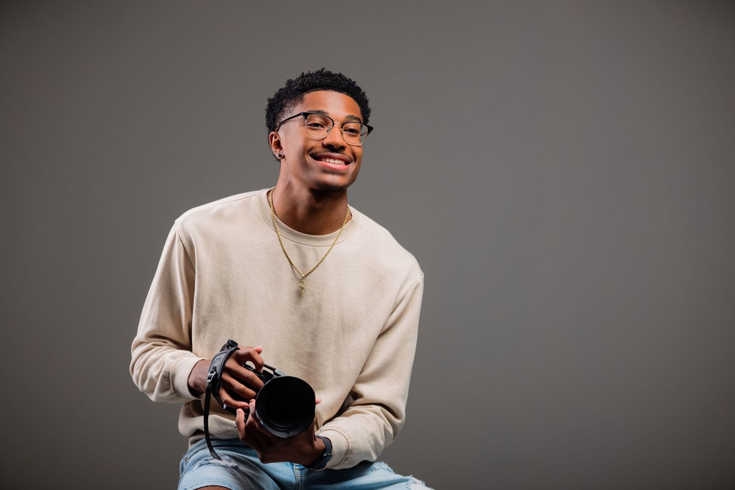 Smiling young man in glasses holding a camera, wearing a beige sweater in a studio setting.