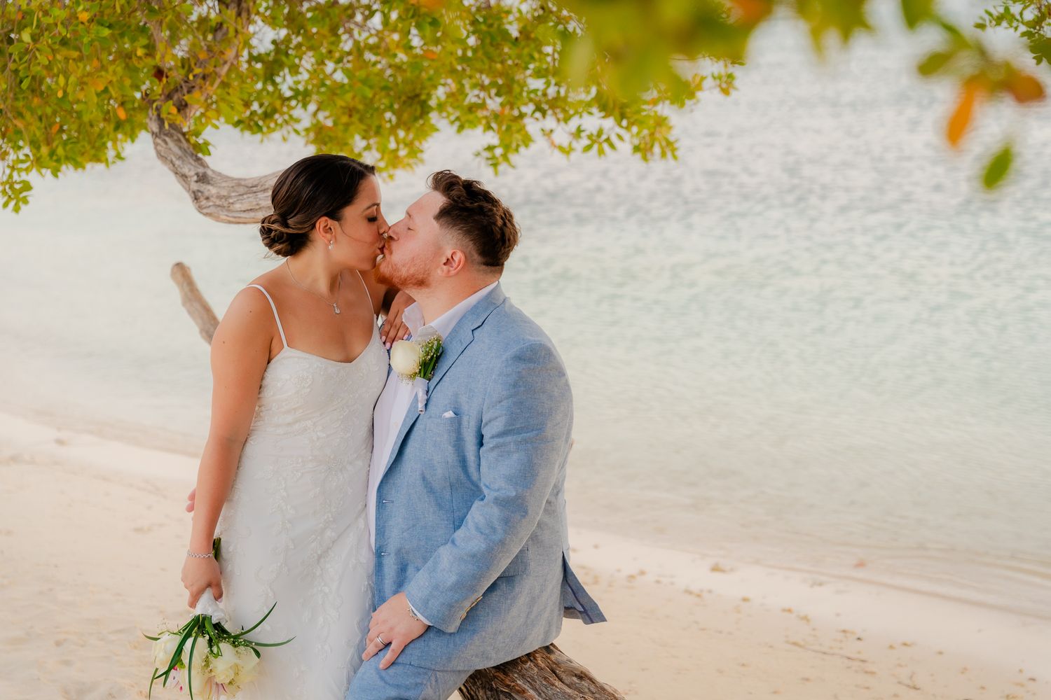 Couple sharing a romantic kiss on a tropical beach under a tree, bride holding bouquet, groom in light blue suit.