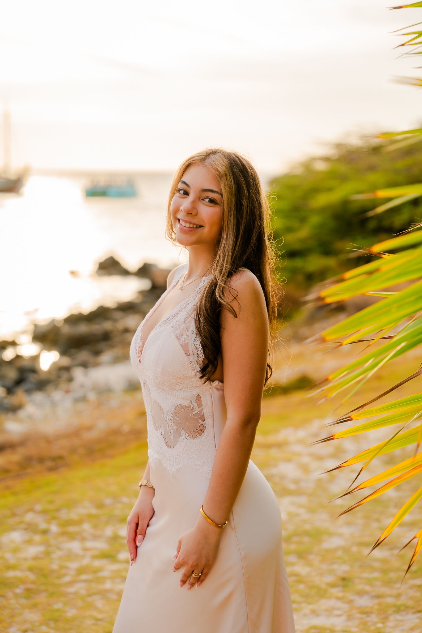 Young woman in white lace dress smiling near palm tree at golden sunset by rocky shoreline.