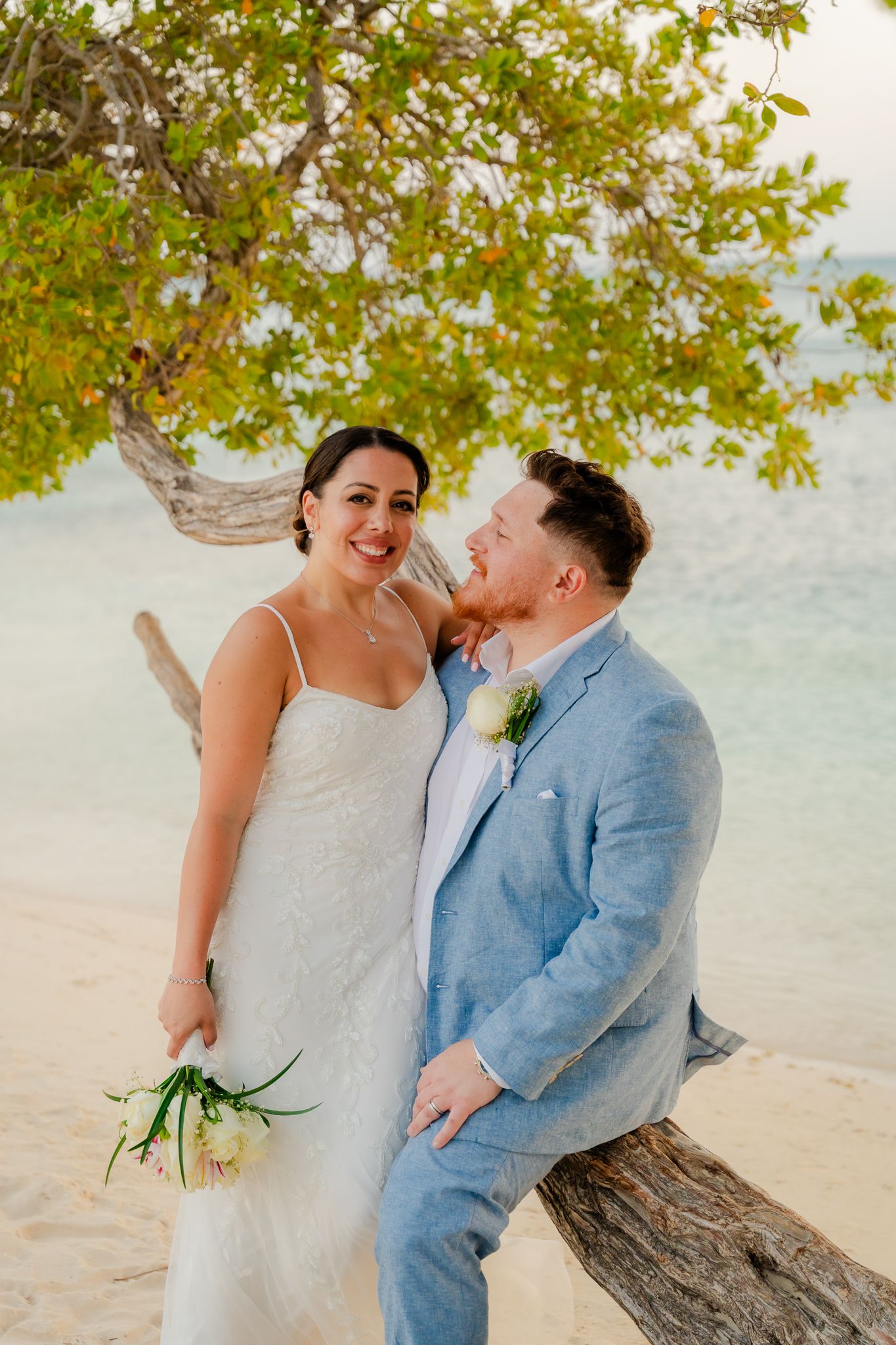 Smiling newlyweds pose under tropical tree on sandy beach, bride holds bouquet in white dress, groom in blue suit.