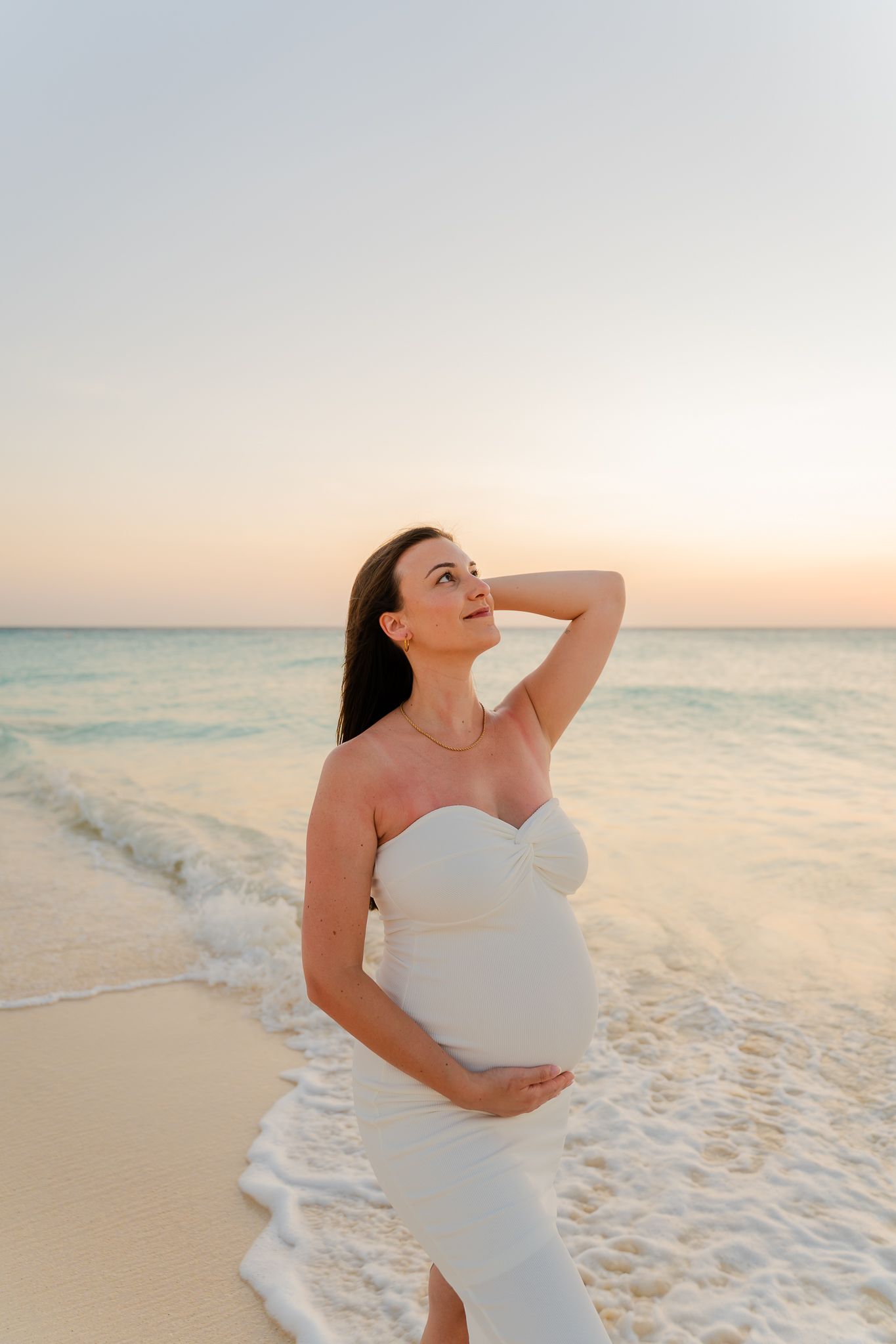 Pregnant woman in white strapless dress standing on beach at sunset, cradling belly and looking up at sky.