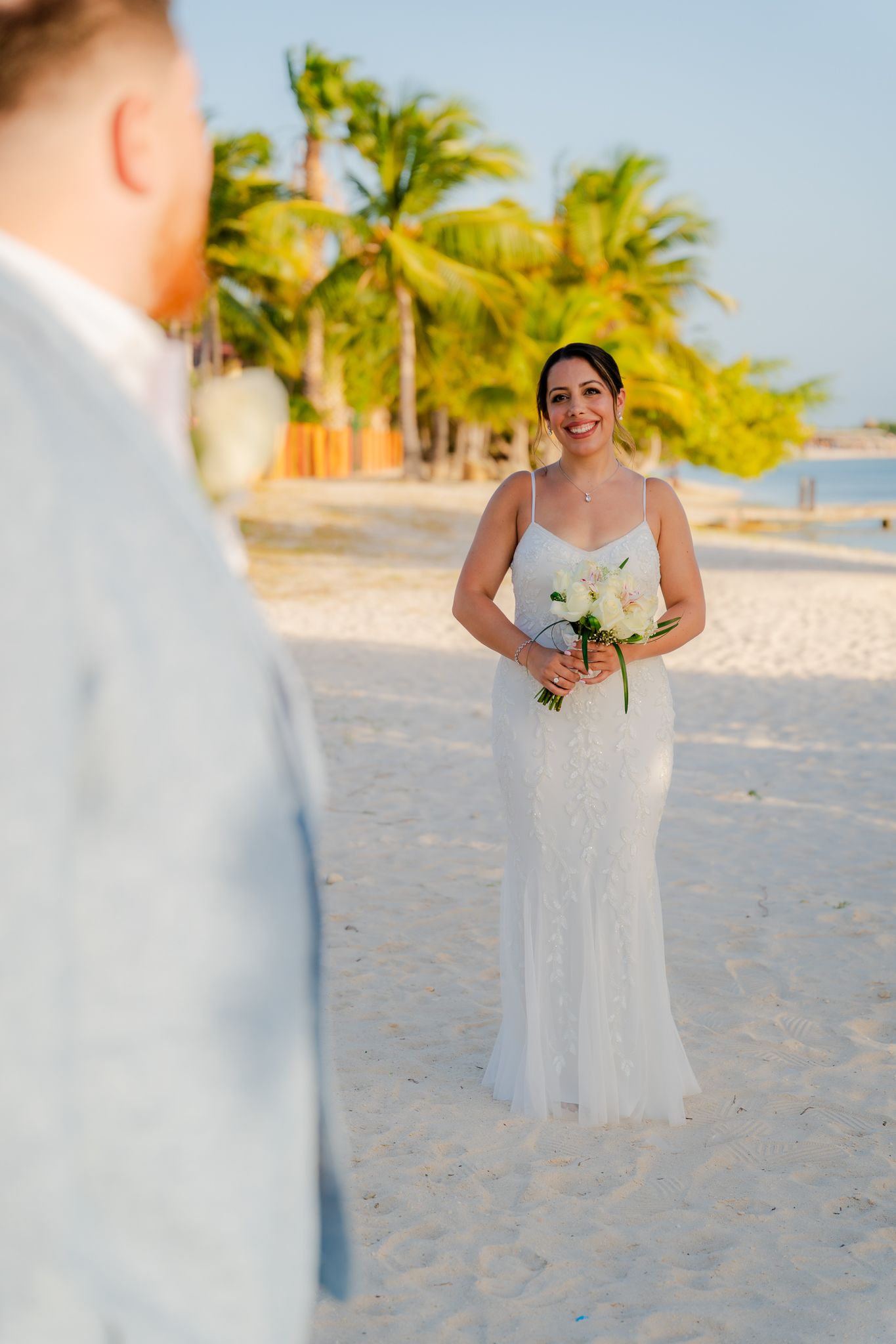 Smiling bride in white gown holds bouquet on sandy beach with palm trees, facing groom in light blue suit.