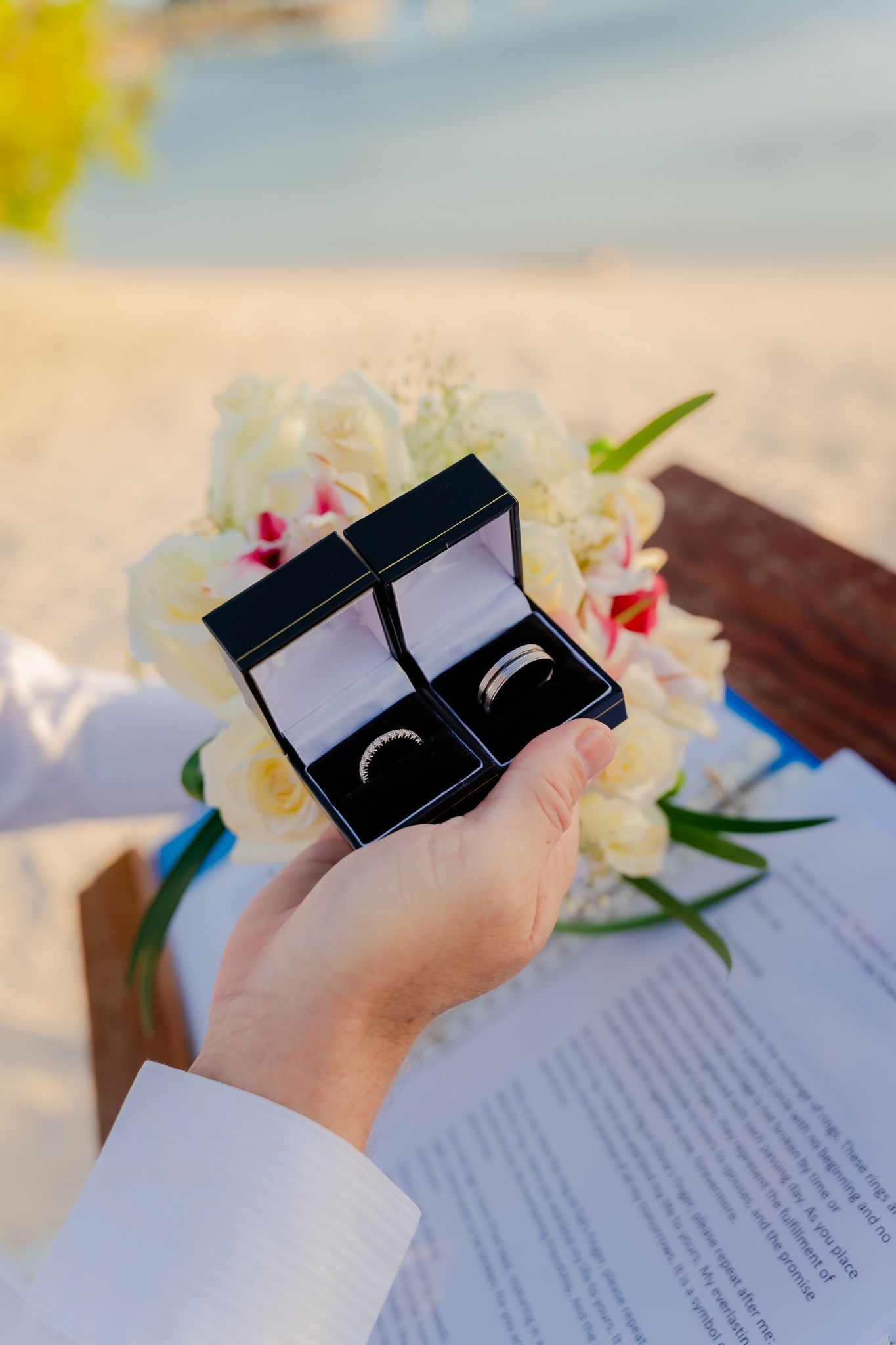 Hand holding two wedding rings in black boxes on a beach with flowers and ceremony documents in background.