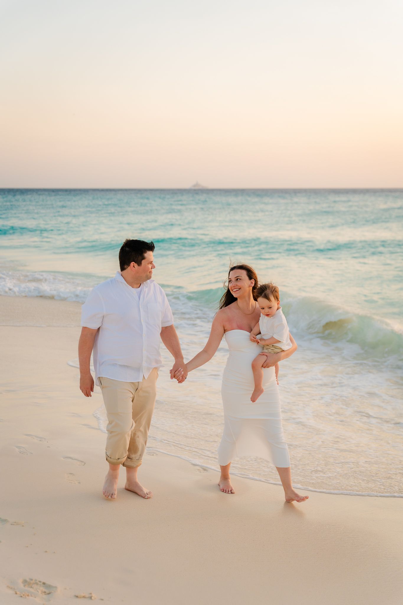 Happy family of three walking on a white sandy beach at sunset, parents holding hands with mother carrying baby.