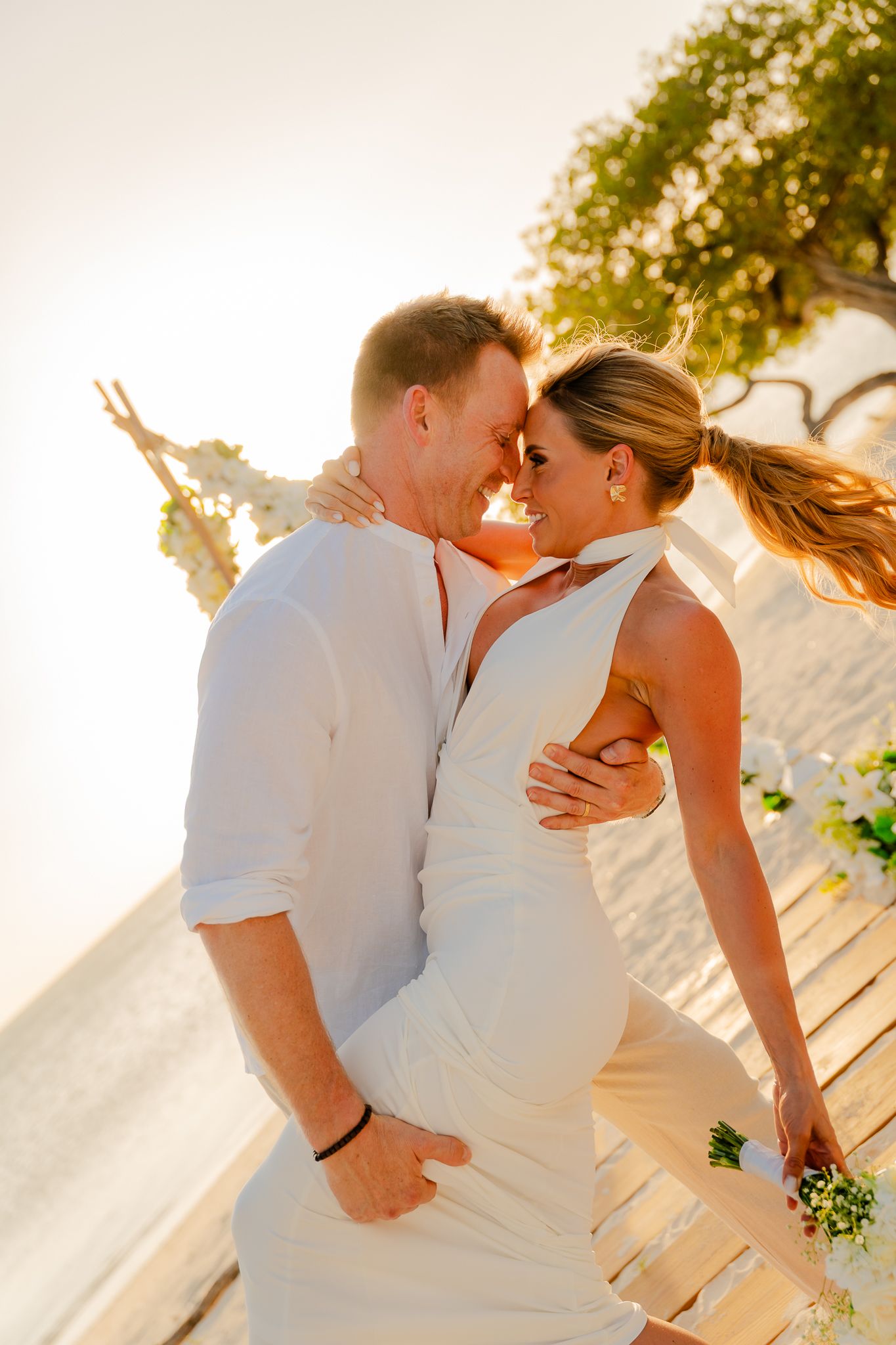 Romantic couple in white outfits sharing an intimate moment at a sunset beach wedding ceremony.
