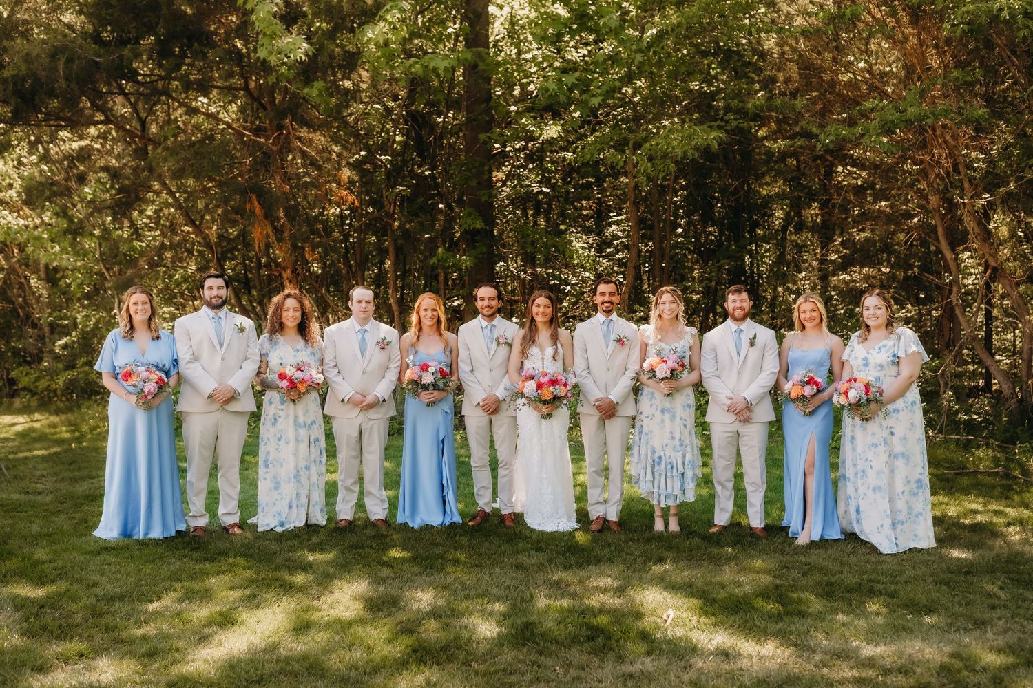 Wedding party posing outdoors in lush greenery, bridesmaids in blue dresses and groomsmen in tan suits flanking the bride and groom.