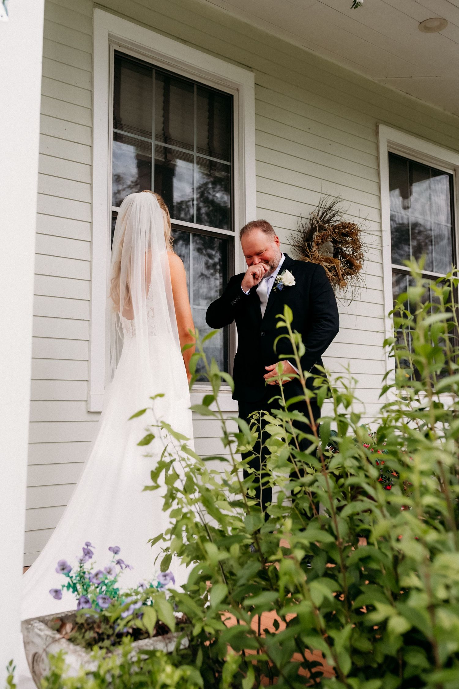 Groom emotional during first look with bride in white wedding gown outside white farmhouse.