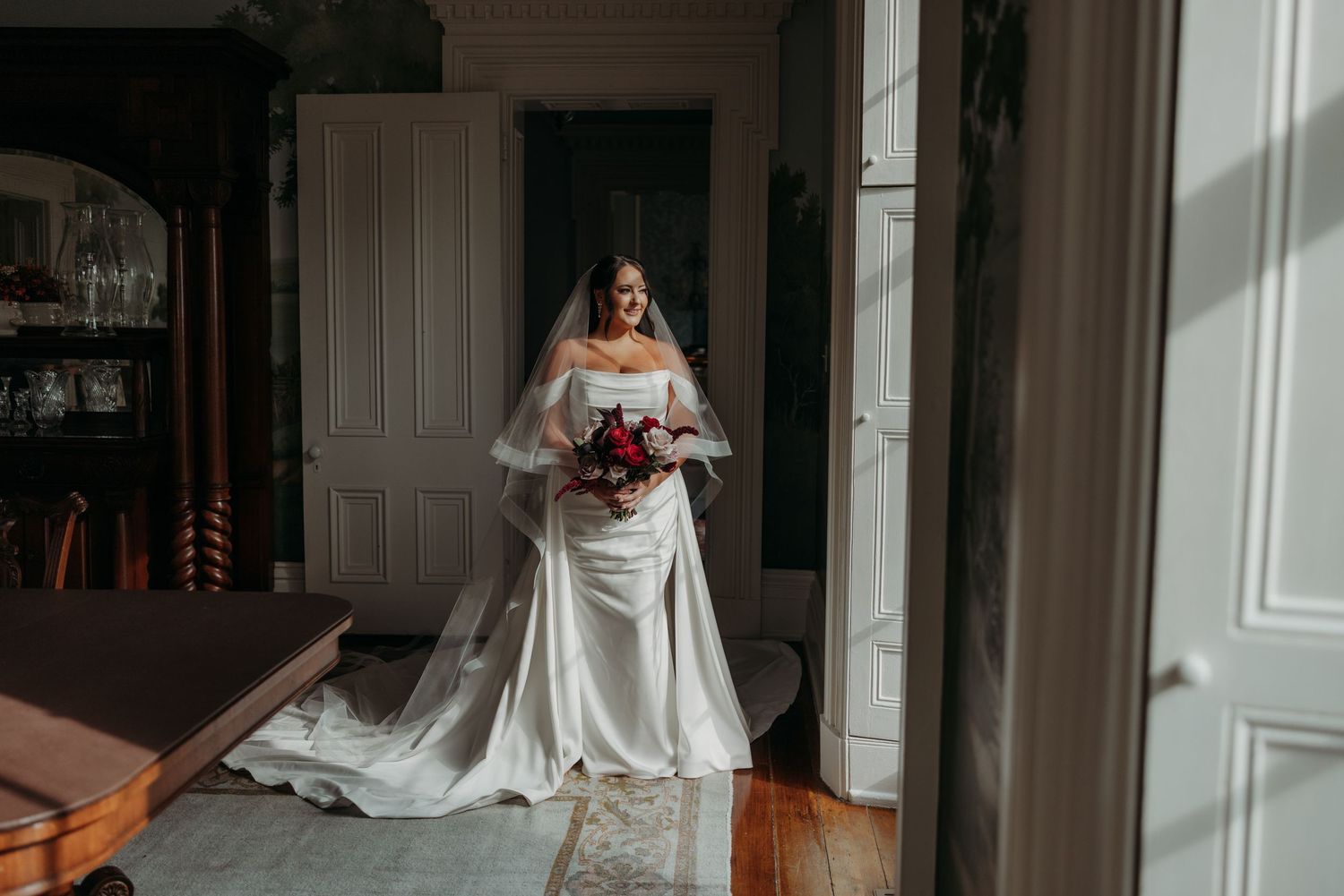 Bride in elegant off-shoulder white gown holding red bouquet stands in sunlit room with long cathedral veil.