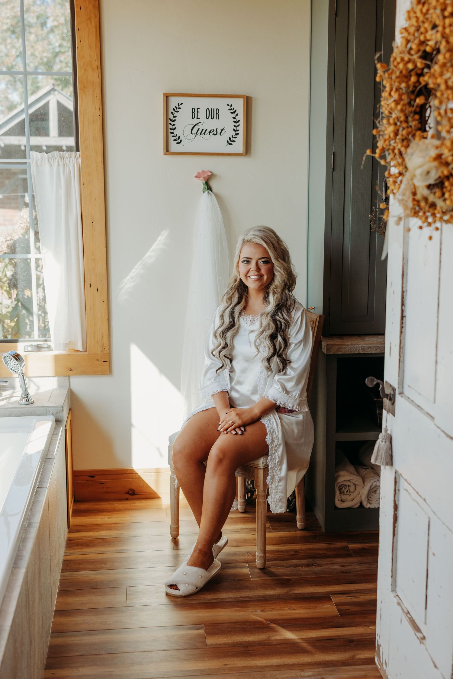 Smiling bride in white robe and slippers sits on wooden chair holding bouquet in cozy rustic bathroom.