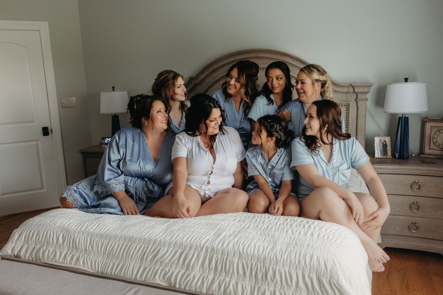 Bride in white pajamas sits on bed laughing with bridesmaids in blue robes during wedding morning preparations.