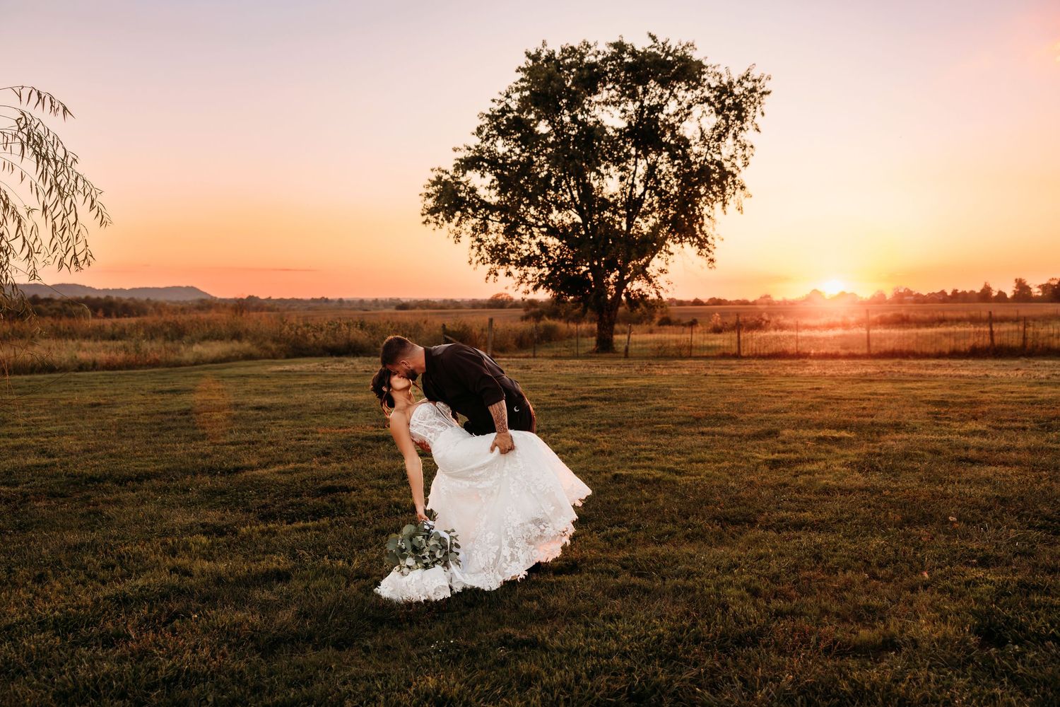 Groom dips bride for romantic kiss at golden sunset in open field during wedding portraits.