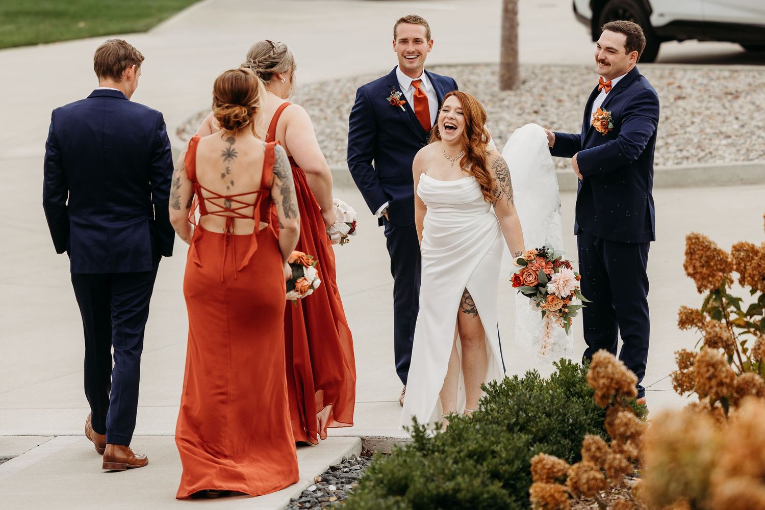 Wedding party in navy suits and rust bridesmaid dresses laughing outside with bride holding bouquet.