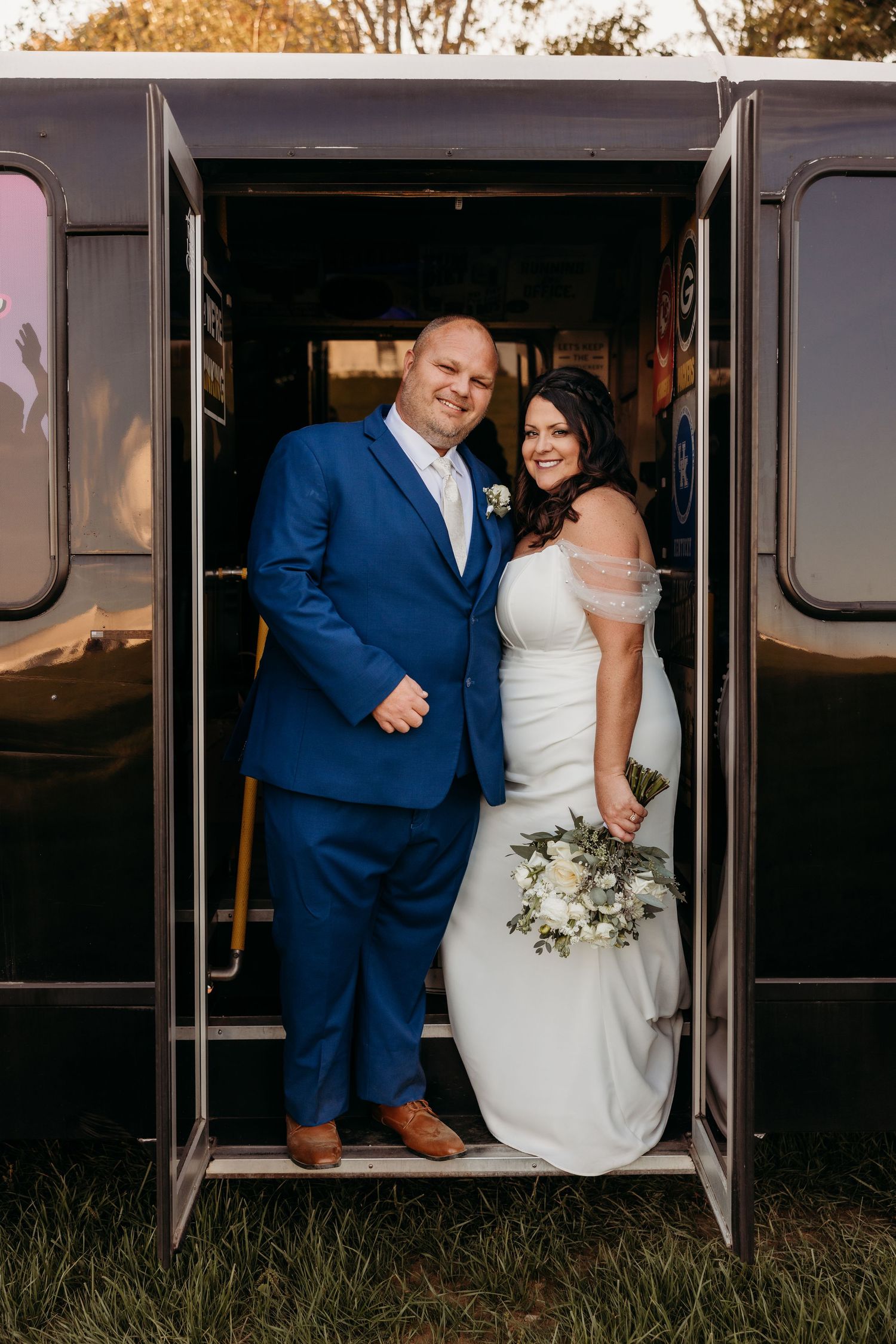 Bride and groom smiling in the doorway of a black party bus, bride holding a white floral bouquet at sunset.