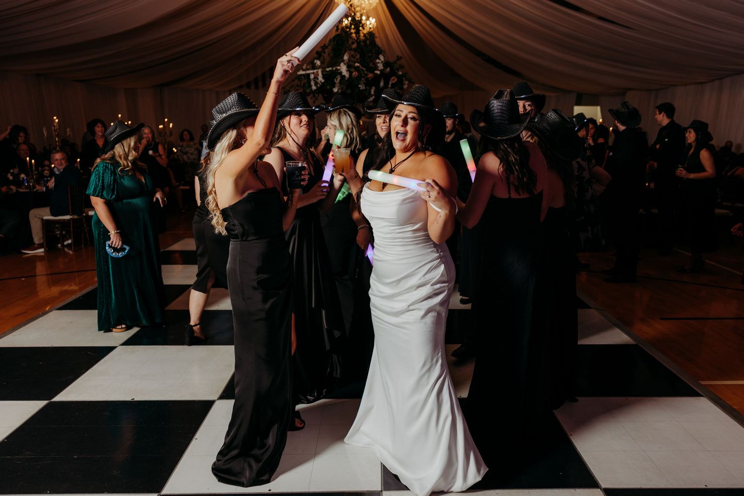 Bride in white fitted gown dancing joyfully with bridesmaids holding glow sticks on checkered dance floor at wedding reception.
