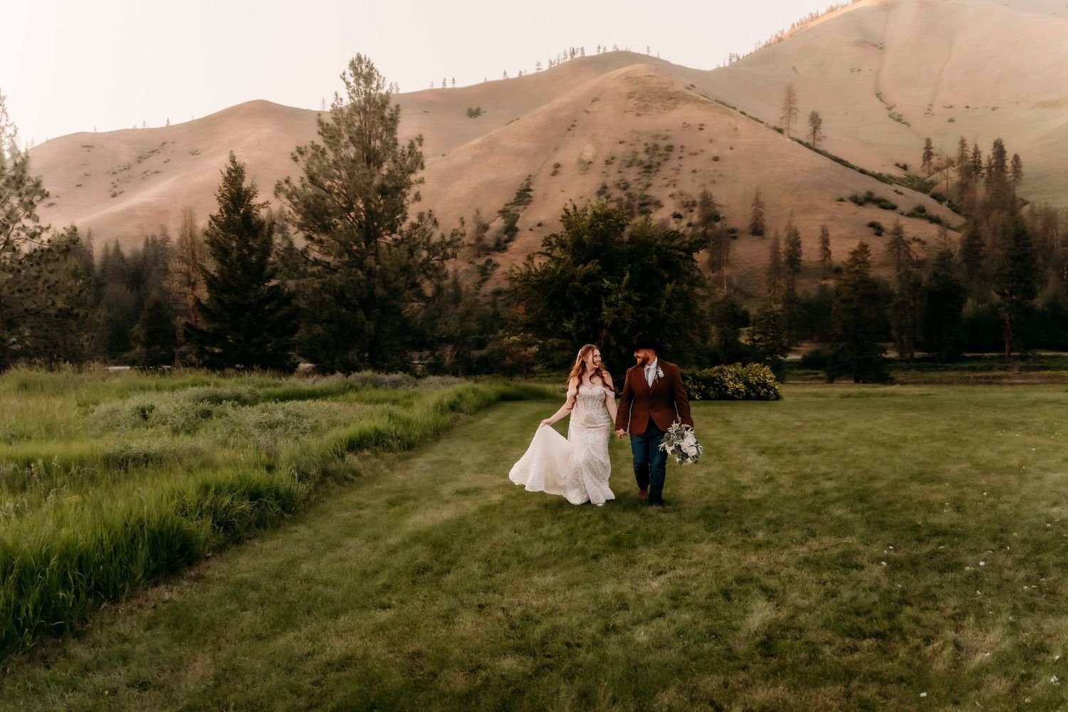 Bride and groom walk hand-in-hand through lush green meadow with stunning mountain scenery at golden hour.