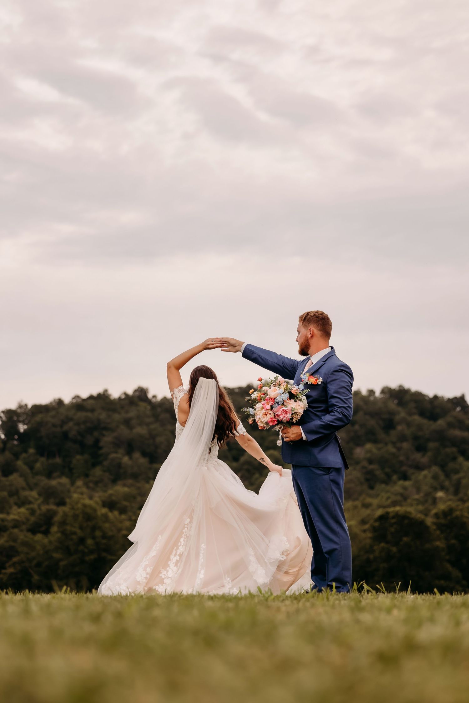 Bride and groom dancing in a green field, groom in blue suit spinning bride in white lace wedding dress with bouquet.