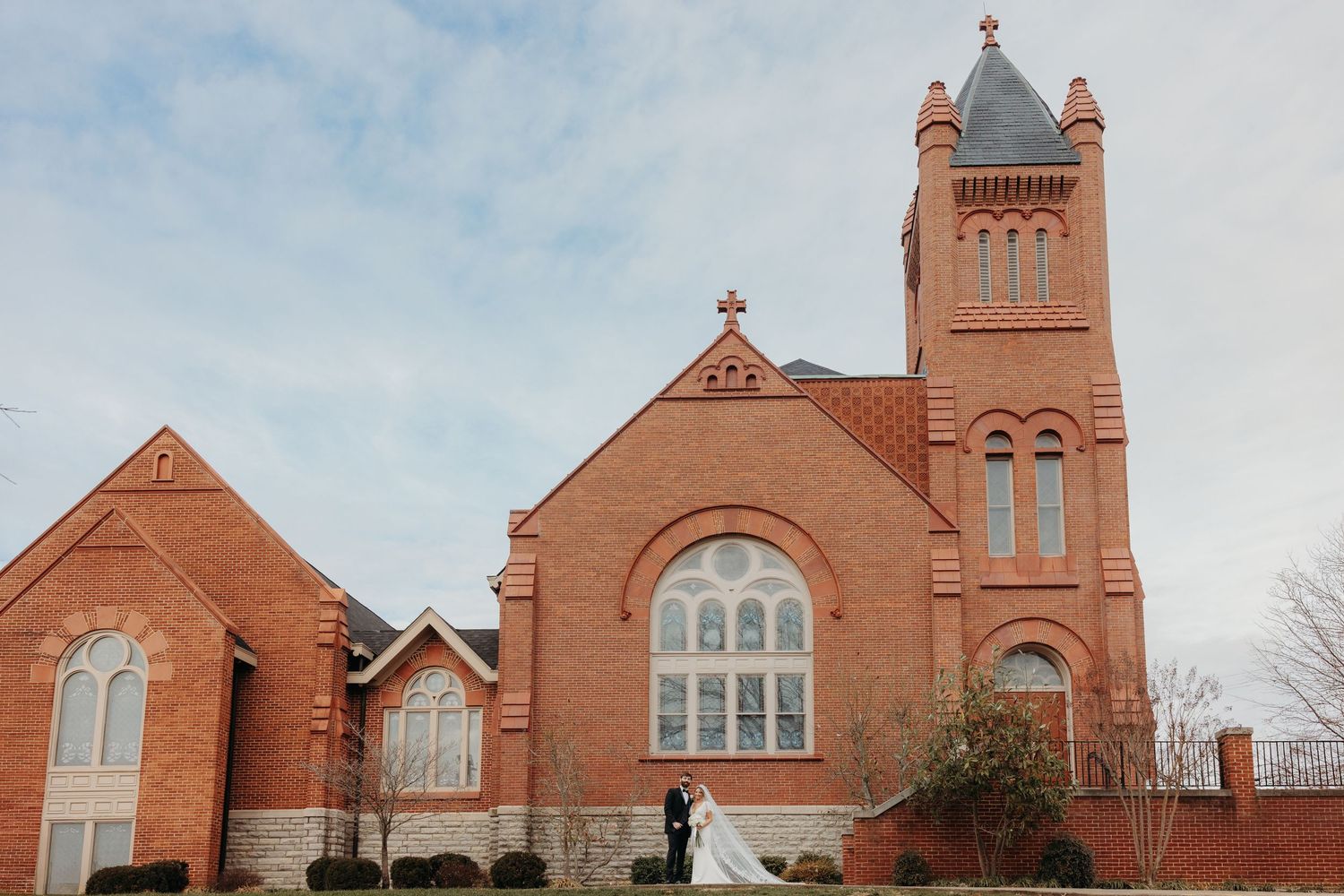 Bride and groom standing outside a historic red brick church with a tall tower on their wedding day.