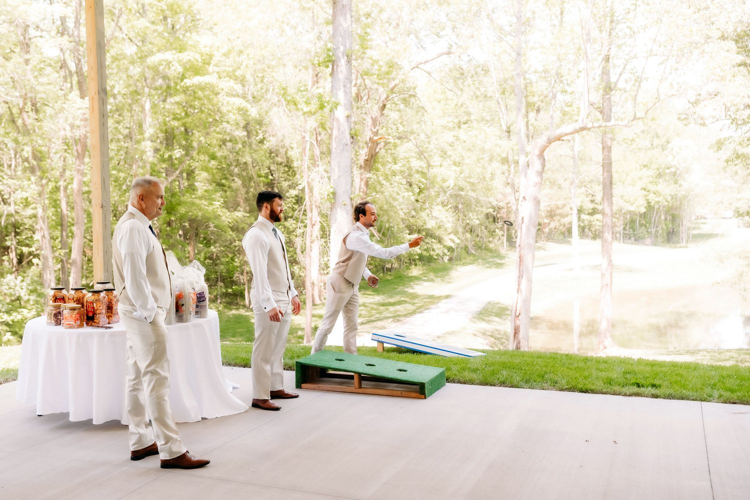 Groomsmen in white suits playing cornhole on outdoor covered patio with trees and pond in background.