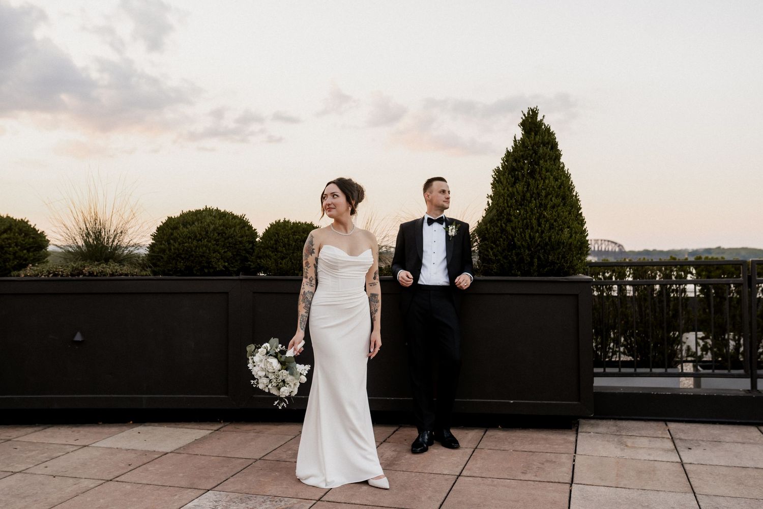 A couple in wedding attire walk together on a rooftop terrace at sunset.