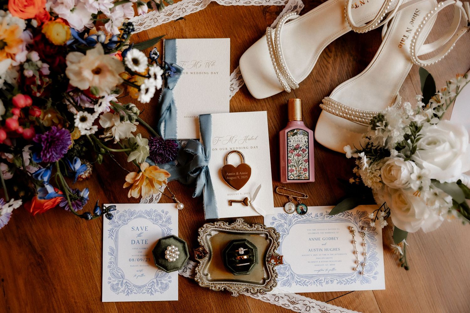 Wedding details flatlay with invitations, rings, and floral elements on wooden surface.
