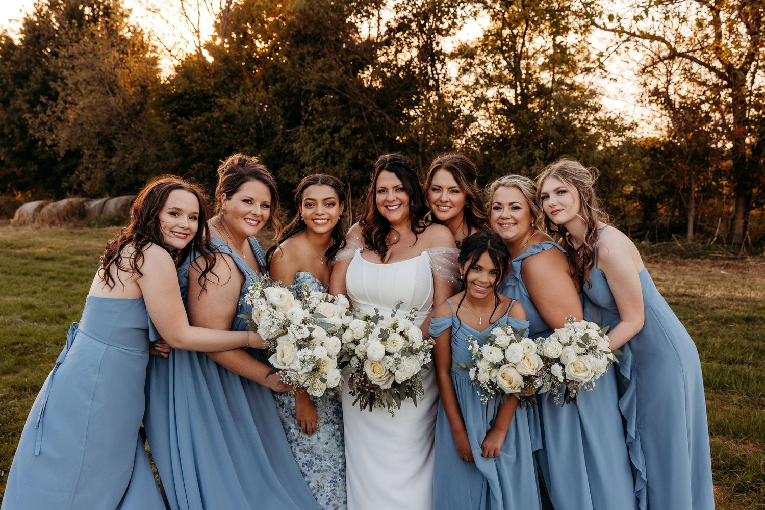 Bridesmaids in powder blue dresses embrace bride while holding white bouquets at sunset.