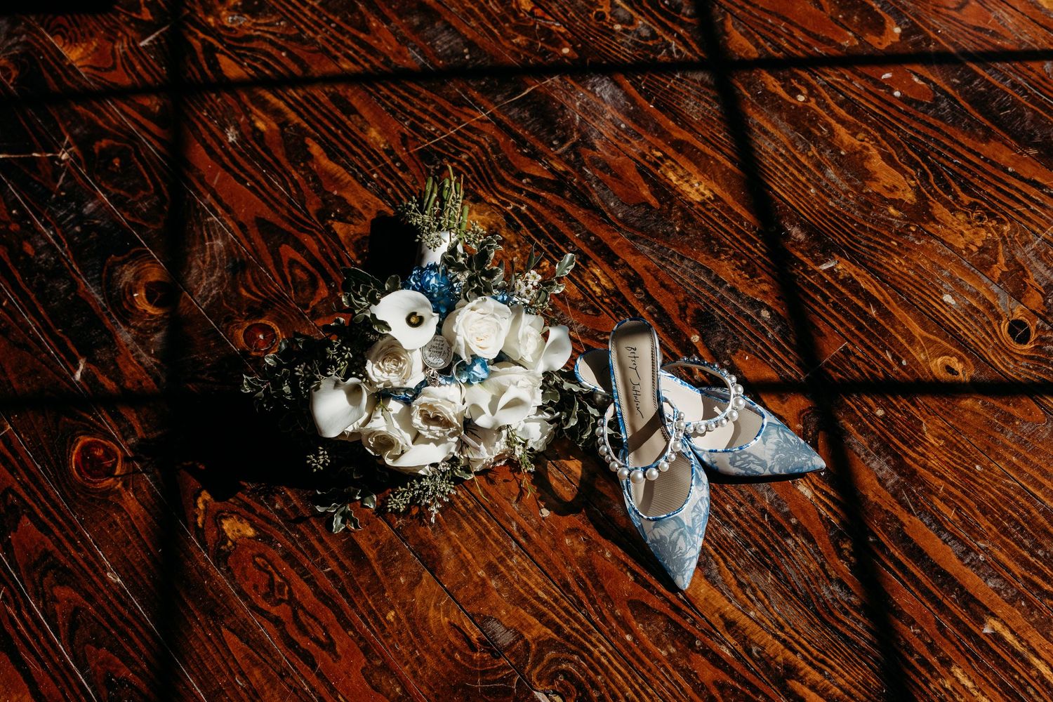 Blue bridal heels and white floral bouquet with greenery arranged on rustic wooden floor in warm sunlight.